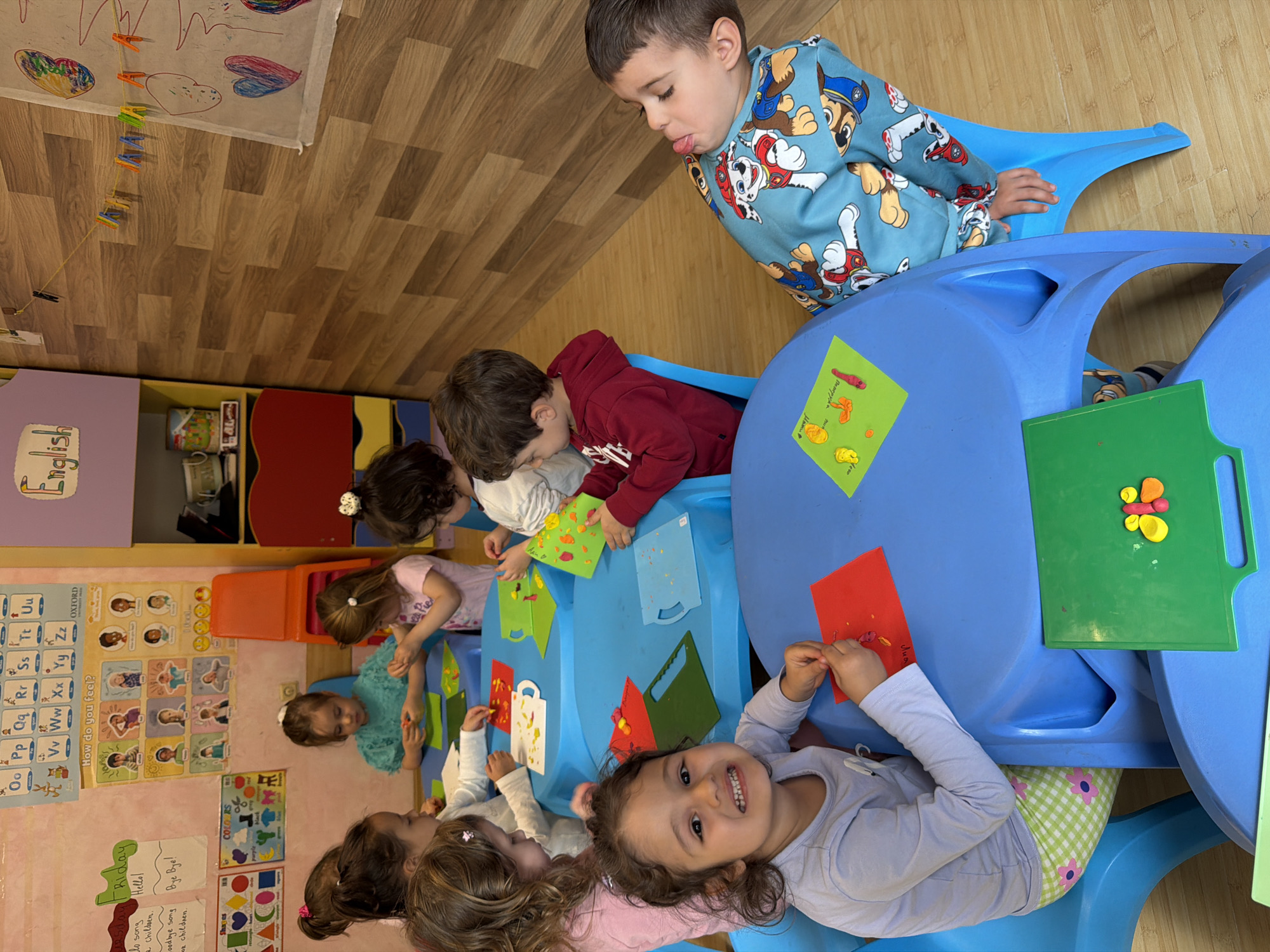 The image shows a group of children sitting around a blue table, engaged in a craft activity with colorful paper and clay. A boy in a Paw Patrol sweatshirt stands beside the table, looking down. The scene appears to be indoors, likely in a classroom or daycare setting, with educational posters and storage shelves visible in the background. The posters include an alphabet chart and "English" label. The environment also contains toys and classroom materials. The children are smiling and interacting with the materials. The location is not explicitly mentioned in the image or text and cannot be determined.