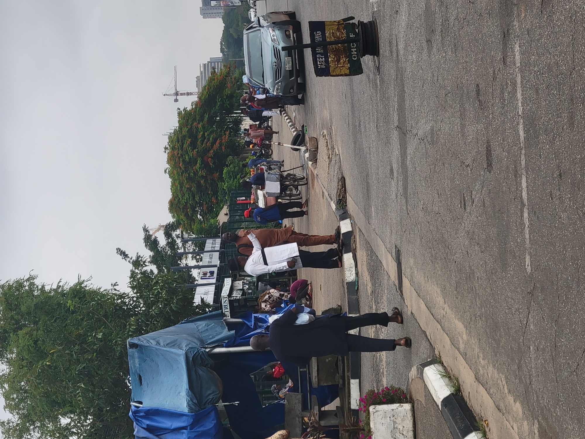 The image captures a street scene in Abuja, Nigeria, with a group of people gathered near a roadside stall. The weather appears overcast, suggesting it might be morning or late afternoon.
Several individuals are visible, some standing and conversing, while others appear to be engaged in transactions or waiting. A blue tarp covers a roadside structure, possibly a stall or shelter. A few bicycles are parked nearby, and a car is visible in the background.
One prominent object is a black and yellow signpost with some text on it. The visible text on the signpost reads "KEEP KING" and "OHCF".
The scene depicts a typical urban street environment with pedestrian activity and roadside commerce. The presence of trees and a faint glimpse of buildings in the background further establish the urban setting. The overall mood seems neutral, depicting a moment of everyday life.