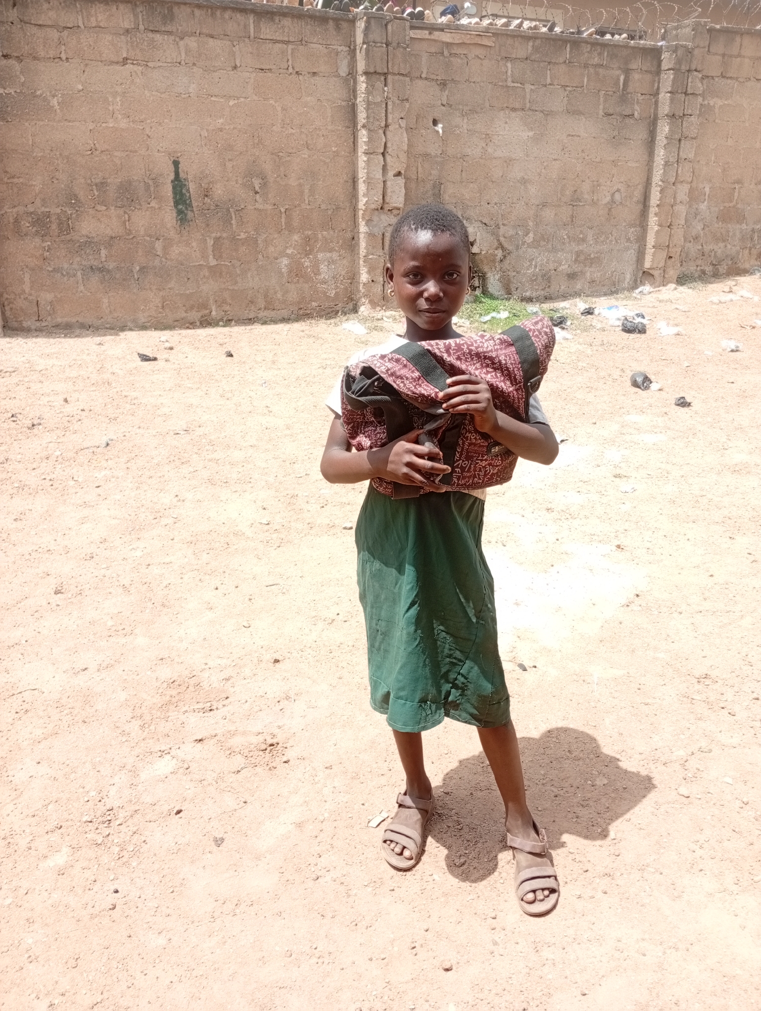 A young African girl stands in a dirt yard, holding a burgundy patterned bag. She wears a green skirt, a light-colored shirt, and sandals. The yard is sandy, and in the background is a brick wall topped with barbed wire. Debris is scattered on the ground. The location is Narayi, Nigeria.