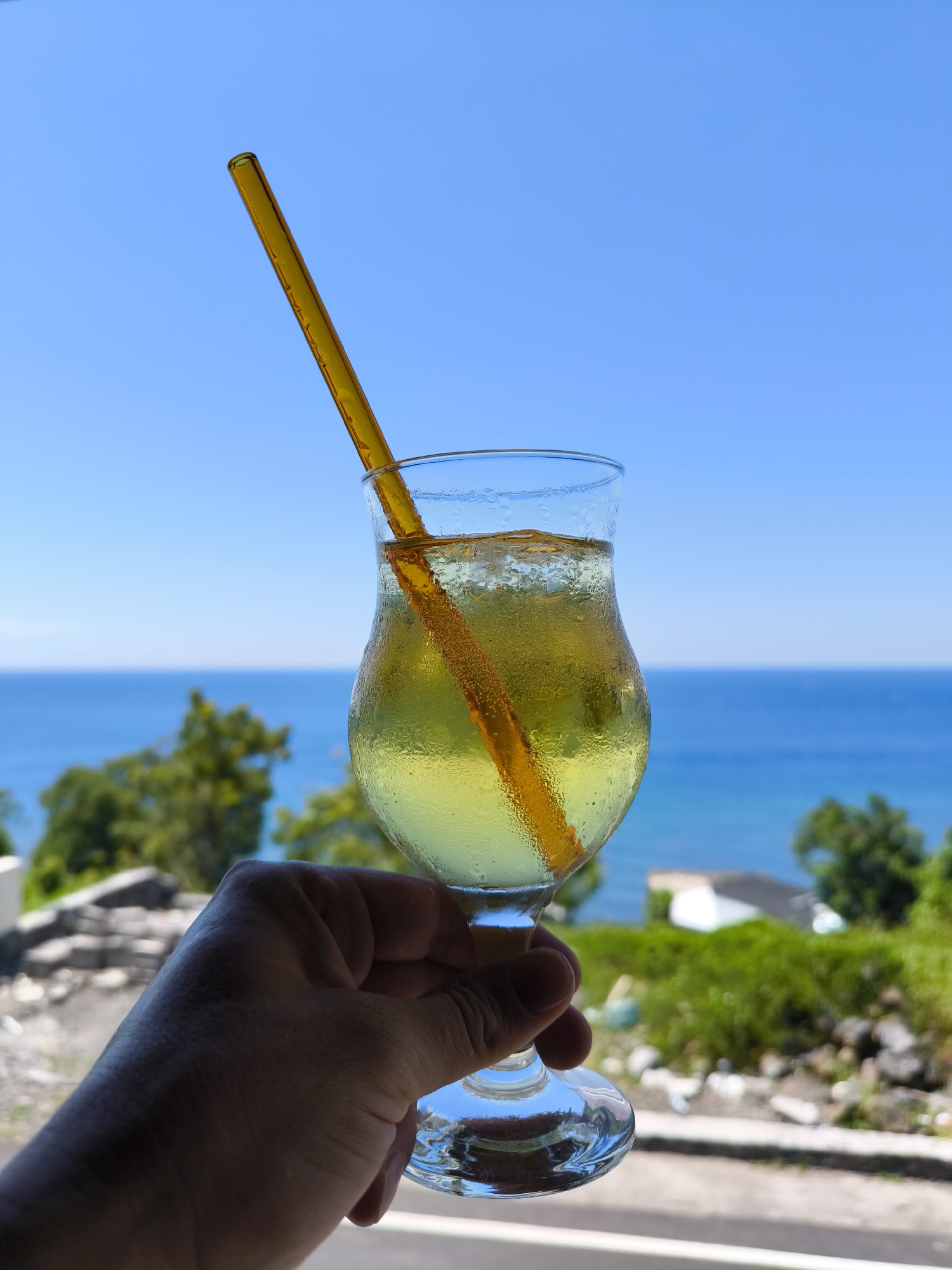 A hand holds a glass of yellow liquid with an amber straw against a backdrop of a clear blue sky, ocean, and green vegetation. The glass is sweating, indicating it is cold. The location is Unknown, Indonesia. Behind the glass, the ocean meets the horizon, and coastal vegetation is visible, including trees and what appears to be a white-roofed building. The environment is sunny and bright. The foreground includes a ledge or surface of some kind.