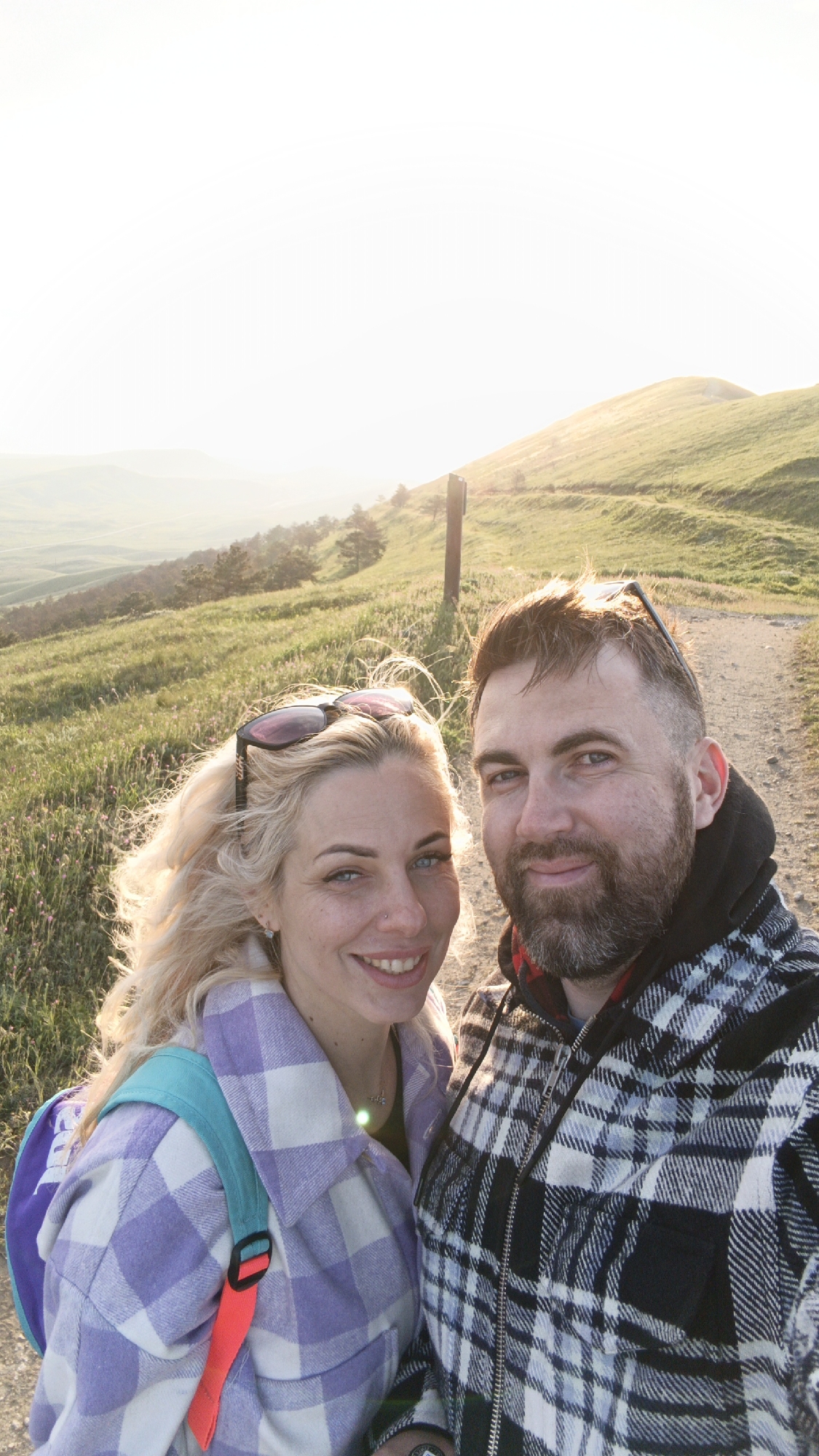 A couple stands on a dirt path in a hilly, grassy landscape. The woman has blonde hair and wears a purple plaid shirt with a backpack. Sunglasses are perched on her head. The man has dark hair and a beard, wearing a black and white plaid jacket over a hooded sweatshirt. A wooden post is visible to their left. In the background are rolling hills under a bright, hazy sky. The location is in a grassy field.