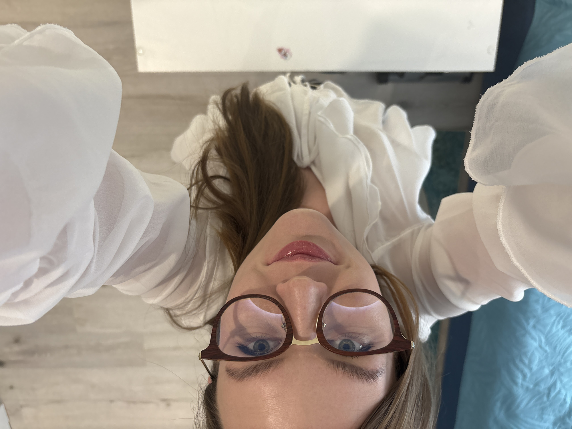 The image is an upside-down, close-up selfie of a person wearing glasses and a white, ruffled blouse. The person has brown hair and fair skin. Their eyes are visible through the glasses. A portion of a white table and a blue quilted object are in the background. There is no indication that the image was taken in Izhevsk, Russia.