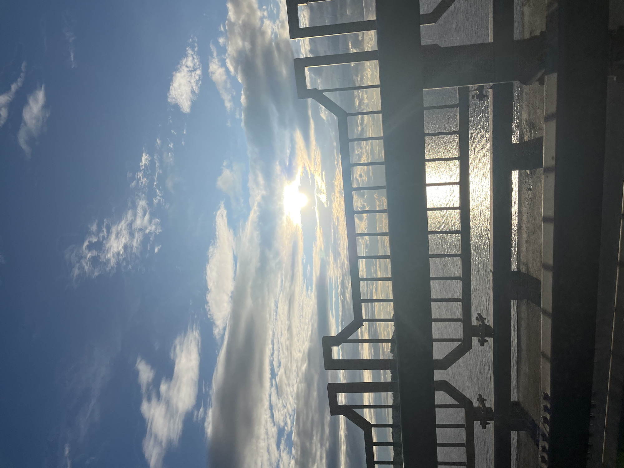 The image shows a view through a metal railing of a body of water with the sun reflecting on its surface. The sky is blue with scattered clouds, and the sun is partially obscured by clouds. The railing is dark and appears to be made of metal. The water is calm, and the reflection of the sun creates a shimmering effect. The scene appears to be outdoors, possibly on a bridge or a pier. The location is identified as Kostroma, Russia. There are no people or animals visible in the image. The overall impression is one of a serene, natural landscape.