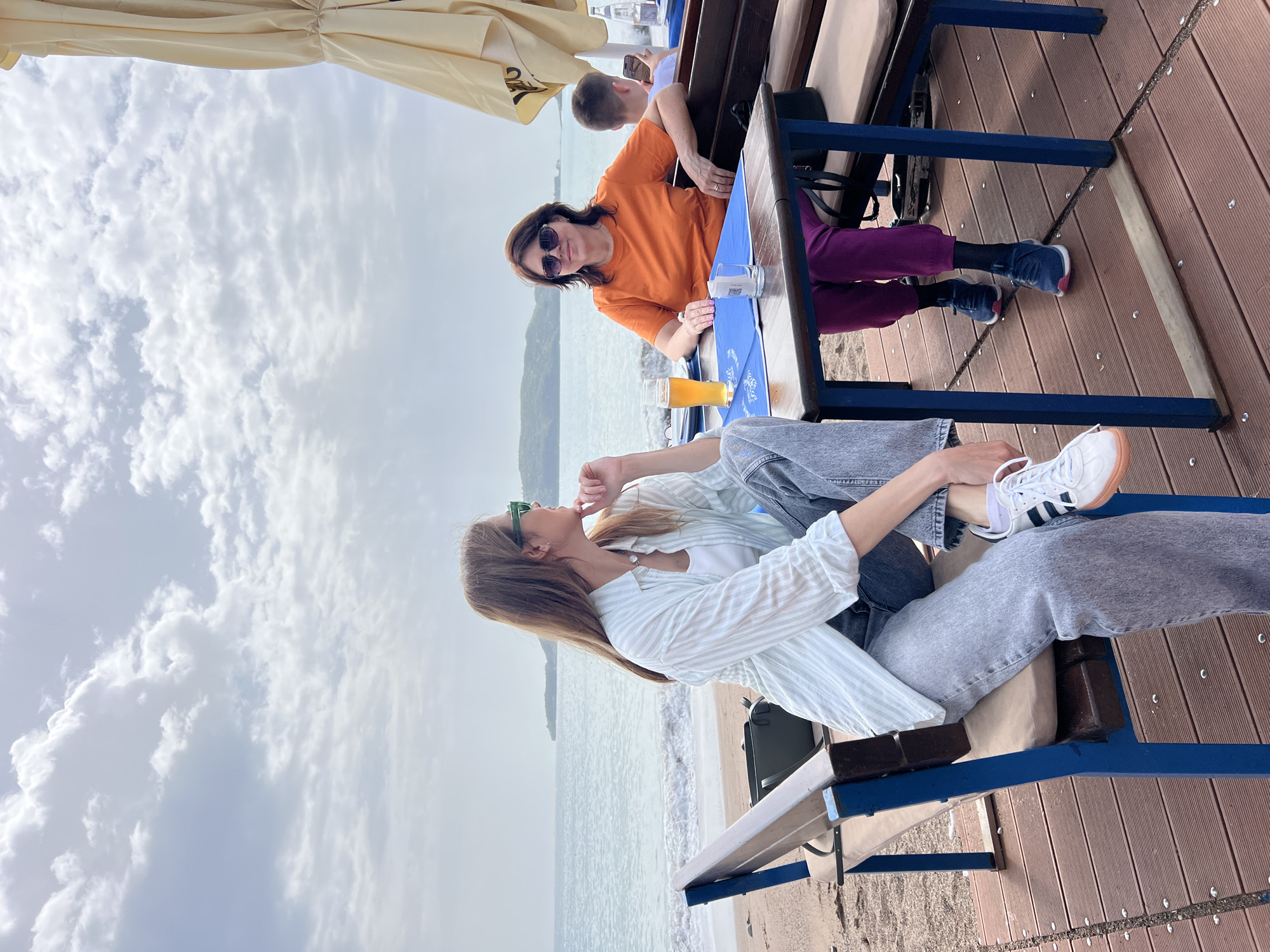 Two women are seated at a table at a beachside restaurant in Budva, Montenegro. The woman in the foreground wears a white striped shirt, light jeans, and white Adidas sneakers with brown soles. She is looking out at the water. The woman behind her wears an orange shirt and purple pants. She is looking directly at the camera and wears sunglasses. A beer glass is on the table between them. The sky is cloudy.
