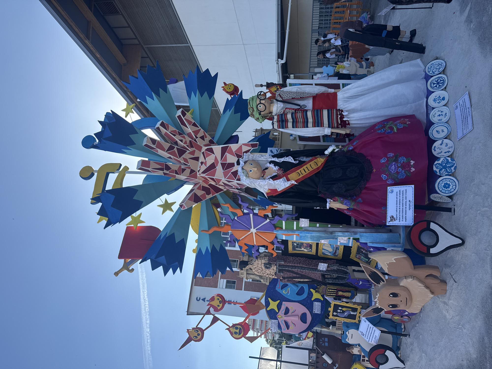 This is a full, eye-level, outdoor shot of a colorful, whimsical sculpture displayed on a sunny day. The sculpture is elaborate, featuring large figures resembling dolls dressed in traditional attire, with a prominent central structure resembling a fan or a burst of stars. The dolls are life-sized or larger, one wearing a black dress with a red sash that reads "BELLEA," and another in a white dress with a striped shawl and green headscarf. Behind the dolls, the sculpture is adorned with various elements like masks, framed pictures, a ship's wheel, and cartoonish figures.
In the background, a brick building with white-trimmed windows is visible, along with a modern building with a gray, corrugated exterior. The sky is clear blue with a few wispy clouds. Several people, including young adults and children, are visible in the background near a fence, seemingly observing the display. The ground appears to be a light-colored, sandy or gravelly surface. The overall impression is of a festive display, likely part of a local event or festival, with a playful and artistic theme.