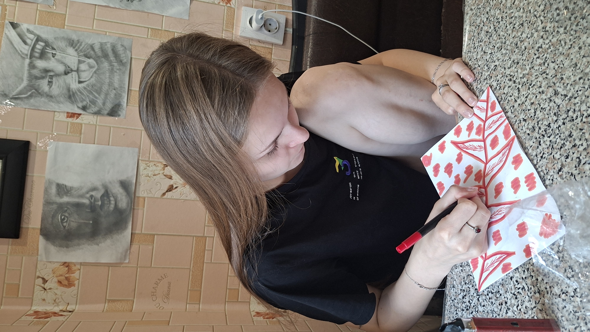 A young woman with long, light brown hair is sitting at a table, drawing on a piece of paper with a red marker. She is wearing a black t-shirt. The drawing appears to be of leaves and small cloud-like shapes, all in red. Her right hand holds the marker, and her left hand rests on the paper. She is wearing a silver ring on her right ring finger. On the table to her left is a red vaping device. The background shows a tiled wall with several framed drawings: one of a crying face, another of a cat, and possibly others partially visible. The environment suggests an indoor setting, possibly a home or a creative space. The overall lighting is moderate.