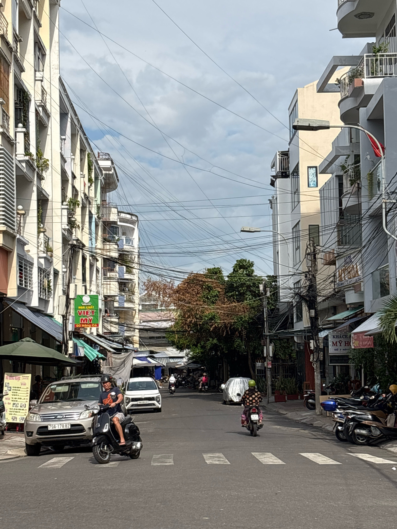 This is a street scene in Nha Trang, Vietnam. In the foreground, a man wearing a dark t-shirt and shorts rides a black scooter. He has a helmet on and is looking forward. He is crossing a pedestrian crossing on a paved road. On the left, a silver SUV is parked with its license plate visible as 79A-178.82. Further down the street, on the right, a woman wearing a patterned shirt and a green helmet is riding a pink scooter. She is also on the road. In the background, there are multi-story buildings on both sides of the street, with balconies and many electrical wires overhead. Trees with brown leaves are visible in the middle distance. Several other scooters are parked on the side of the road. The sky is cloudy with patches of blue. A red flag is visible on a building on the right. Signs in Vietnamese are present on some buildings.