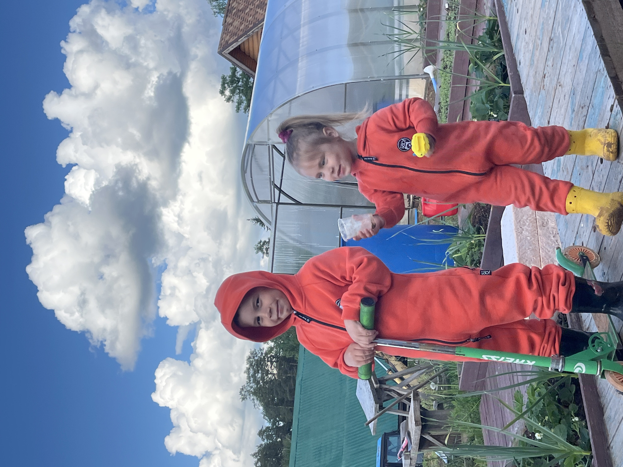 The image shows two children in a backyard or garden setting. Both children are wearing orange jumpsuits. The child in the foreground is wearing black boots and holding a green scooter. The child in the background is wearing yellow boots and holding a yellow toy and a clear plastic cup. Behind them is a greenhouse-like structure. The sky is visible, with blue sky and white clouds. It is unclear from the image if the location is in Chaikovsky, Russia.