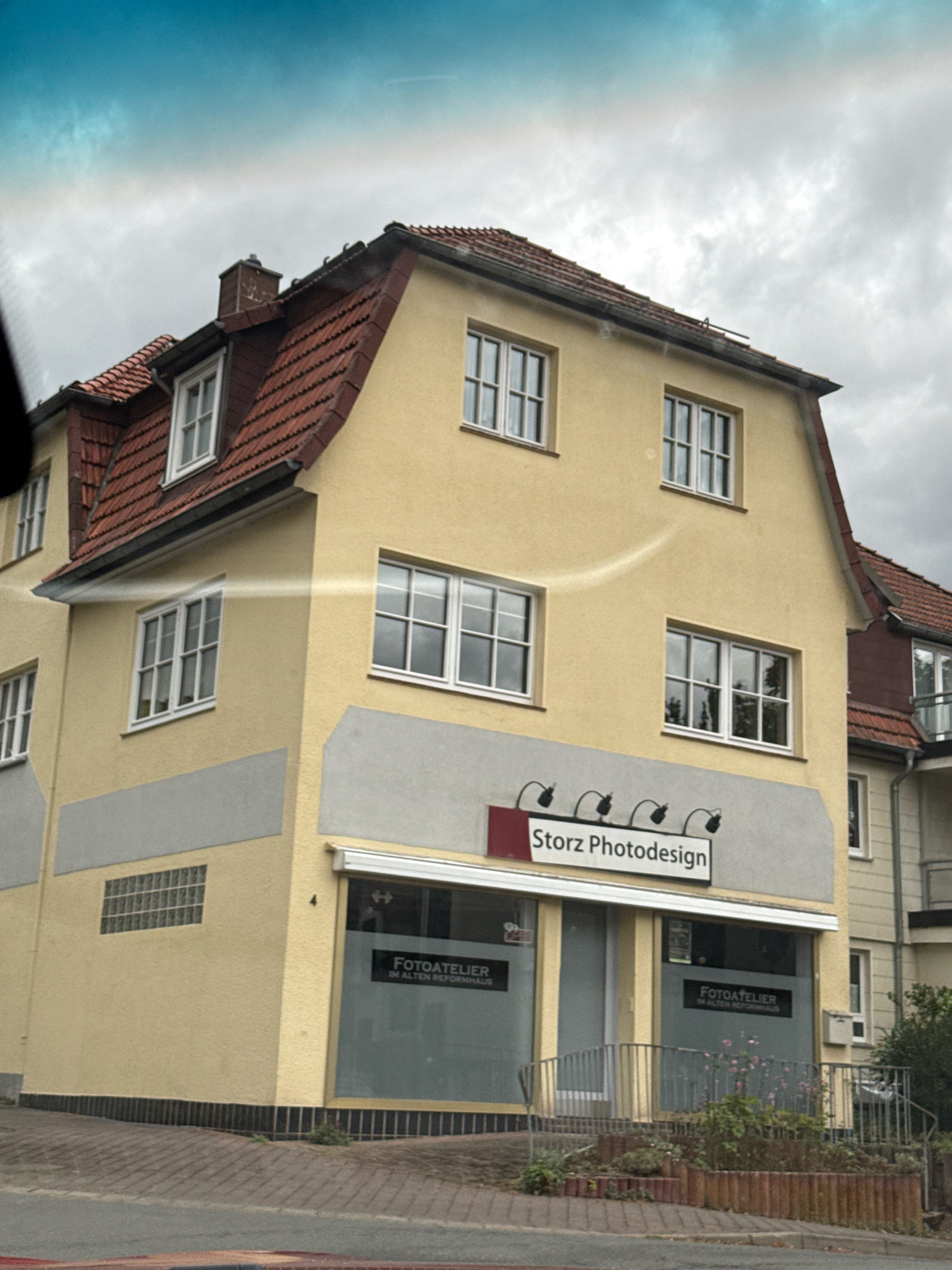 The image shows a two-story yellow building with a red tiled roof in Bad Sachsa, Germany. The building has several windows on both floors. The ground floor has a business with signage that reads "Storz Photodesign" and "FOTOATELIER IM ALTEN REFORMHAUS". There is a ramp leading to the entrance of the business, with a metal railing and some plants. The building appears to be part of a larger complex, with another building visible to the right. The sky is overcast.
