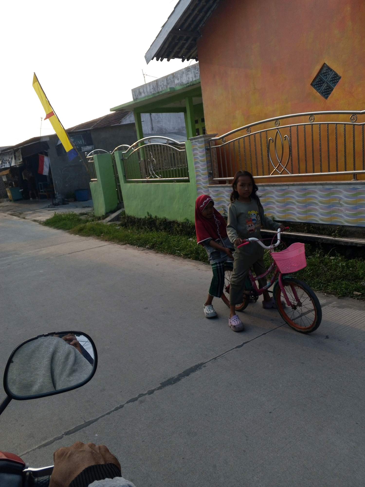 Two young girls are riding a pink bicycle on a paved road in Ciruas, Indonesia. The girl in front is wearing a long-sleeved gray shirt with a cartoon print and dark pants. She is pedaling the bicycle. The girl behind her is wearing a red hijab and a gray and green striped garment. She is seated on the back of the bicycle. The bicycle has a pink basket attached to the handlebars. The scene appears to be in the late afternoon, with the sun casting shadows. In the background, there is a yellow flag on a pole, a green fence, and an orange building with a diamond-shaped window. The road is made of asphalt and has a visible crack running down the middle. A motorcycle's side mirror and a hand are visible in the foreground, suggesting the photo was taken from a motorcycle.