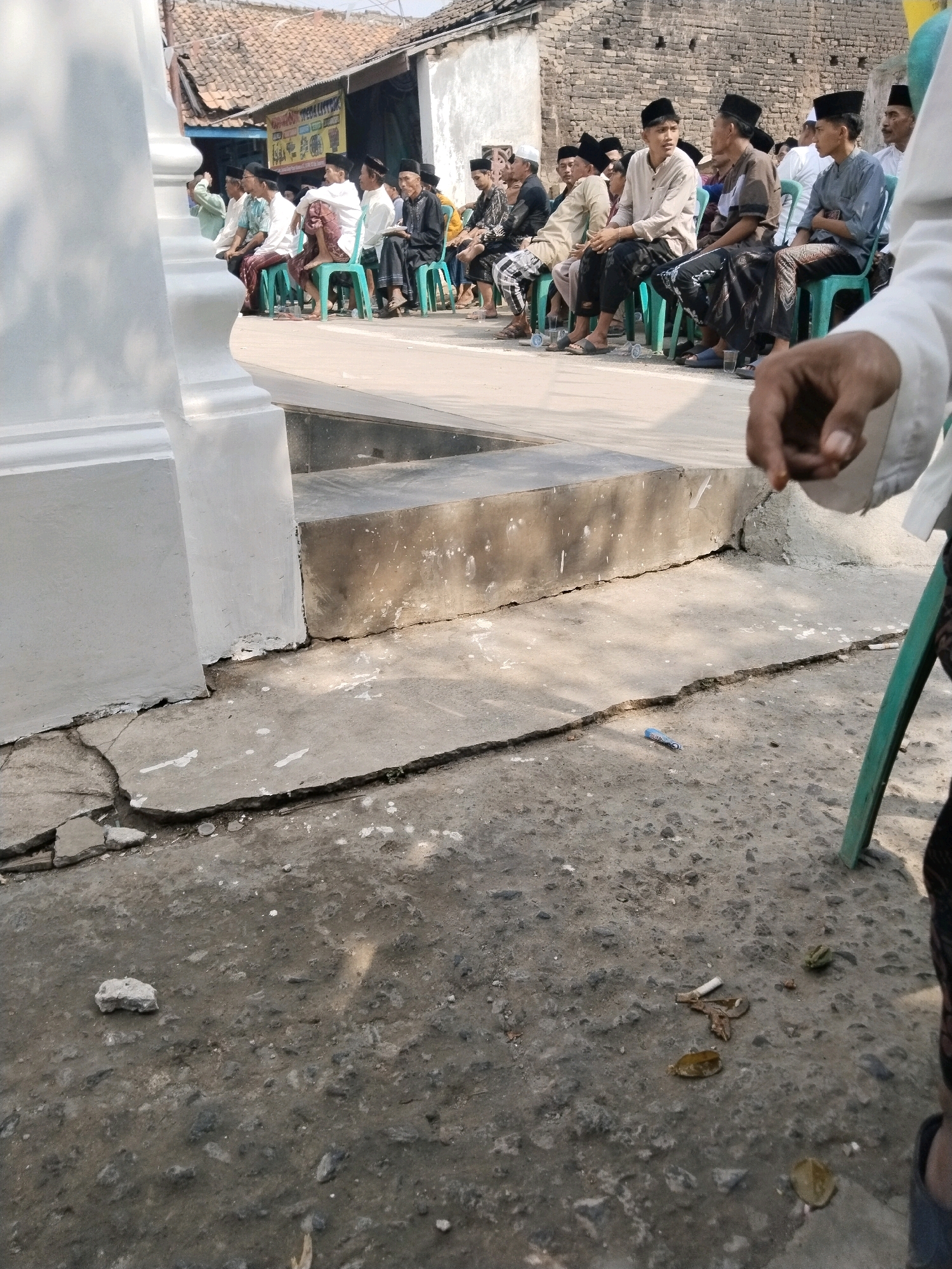 In Serang, Indonesia, a group of men are seated on green plastic chairs arranged in rows. They are all wearing traditional Indonesian attire, including peci hats and sarongs. The men are facing in the same direction, suggesting they are attending an event or ceremony. The background shows a brick wall and a building with a tiled roof, typical of Indonesian architecture. The ground in the foreground is cracked concrete, with some debris scattered around. The lighting suggests it is a sunny day.