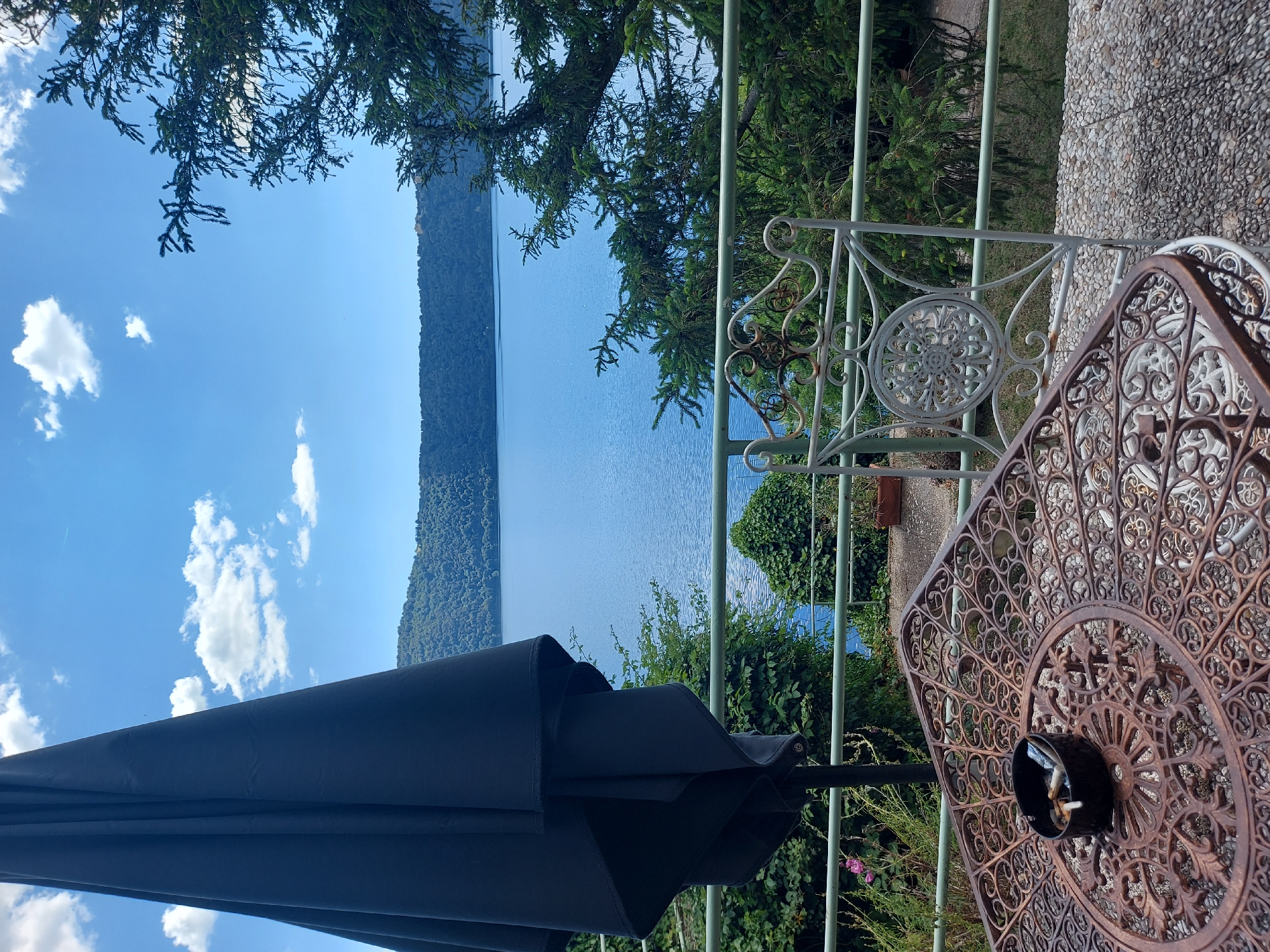From a high vantage point, a photograph captures a scene likely in Castel Gandolfo, Italy, overlooking Lake Albano on a clear day.
The foreground features a patio setting. A decorative wrought-iron table, primarily brown in color with an intricate patterned surface, is partially visible on the right side of the frame. An ashtray containing cigarette butts sits on the table. Next to it is a white, wrought-iron chair with a circular back design. A dark-colored patio umbrella provides shade, occupying the lower left of the frame.
A white railing partially obscures the view of Lake Albano, which stretches horizontally across the midground. Dense green forests line the distant shore, contrasting with the clear blue sky and water. A few white clouds are scattered across the sky. The branches of trees frame the upper portion of the image, adding a natural element to the scene.