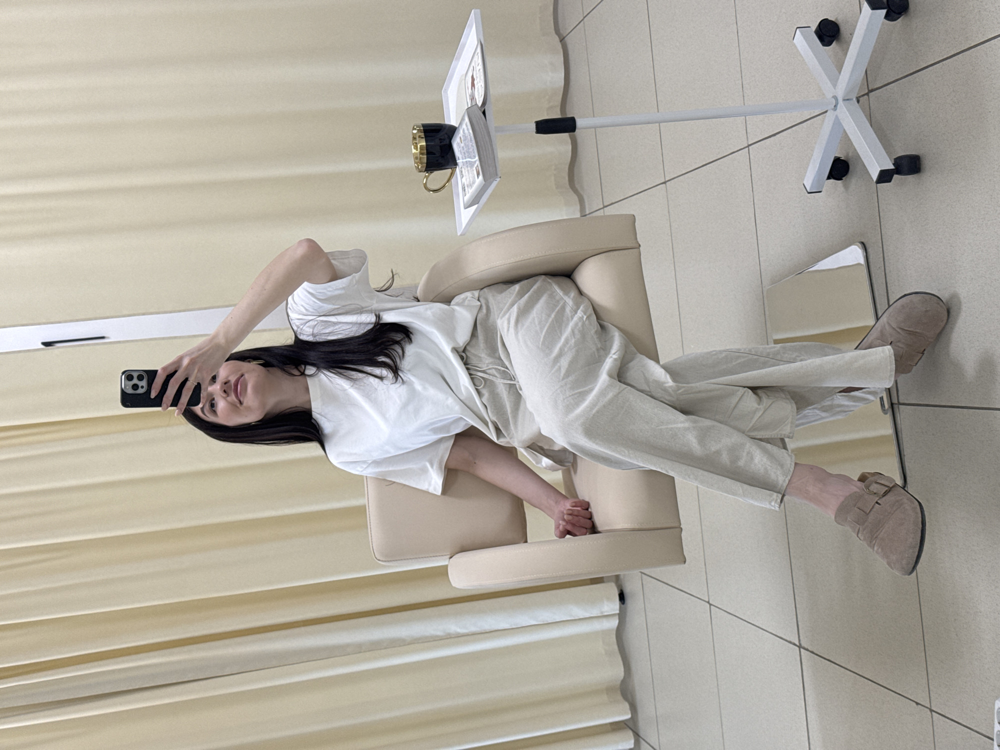 An indoor scene in Chaikovsky, Russia, depicts a female seated in a light beige upholstered armchair. The subject, with long dark hair, is wearing a white short-sleeved t-shirt, light beige wide-leg pants with a visible drawstring, and light brown suede mule-style shoes with a buckle. She holds a black smartphone, identifiable by its triple rear camera module, in her raised right hand, pointing it downwards as if taking a photo or video. Her gaze is directed upwards towards the phone. Her left hand rests on the armrest of the chair.
To the subject's right, a white height-adjustable mobile side table stands on a cruciform base with black caster wheels. On the tabletop, a black mug with a gold interior and handle is positioned to the left of an open book or magazine. The floor consists of large square light-colored ceramic tiles with dark grout lines. The background is dominated by light beige vertical curtains, providing a soft, uniform backdrop. A vertical white reflective surface, possibly a mirror or part of a partition, is visible behind the subject to the left.