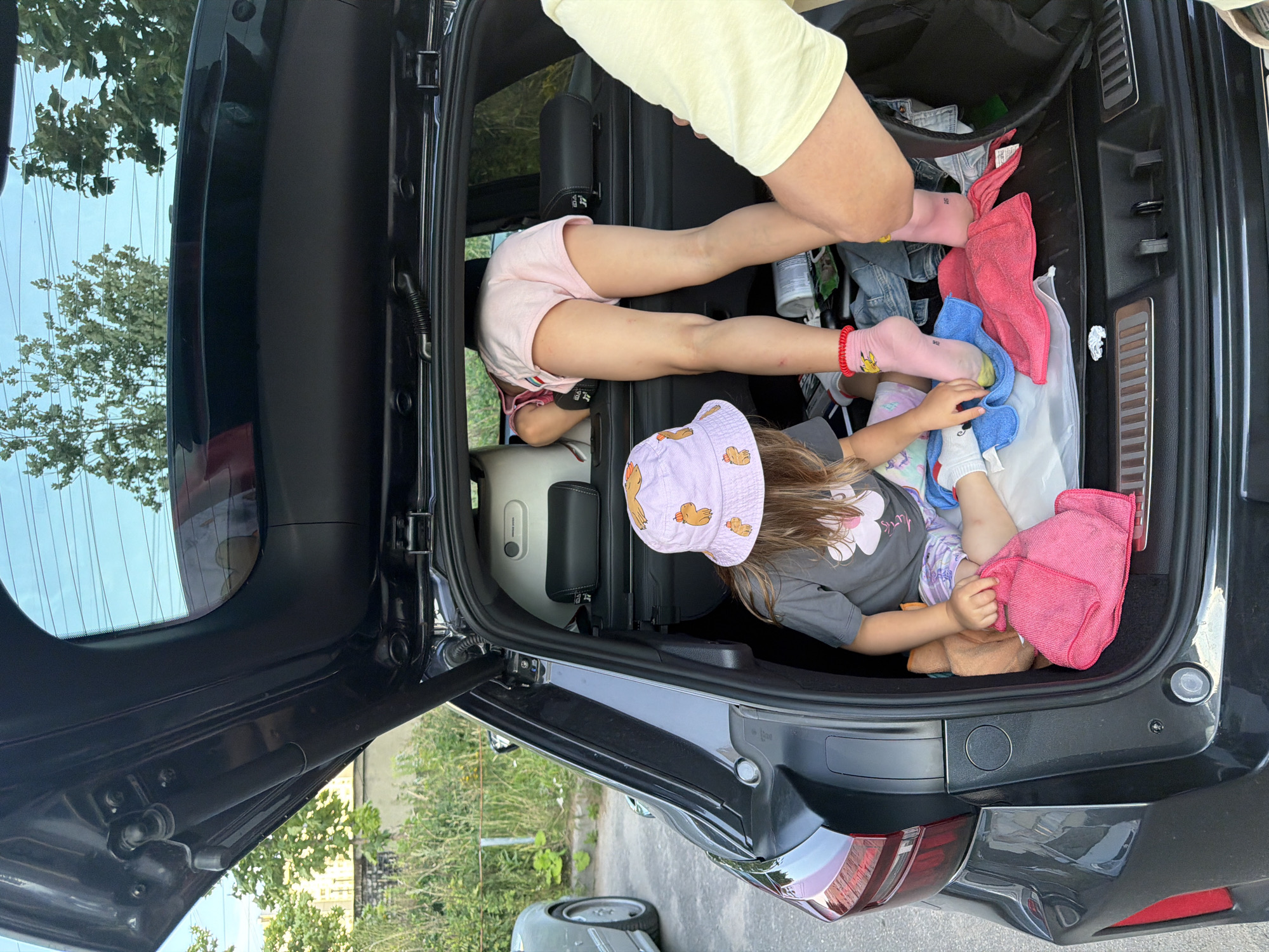 In Andreyevka, Russia, two young girls are observed rummaging through the open trunk of a dark-colored SUV. The girl in the foreground, wearing a purple bucket hat with cartoon characters and a grey t-shirt, is sitting and holding a pink towel. The second girl, dressed in pink shorts and a t-shirt, is bent over with her upper body inside the trunk, seemingly searching for something. Her legs are visible, one with a pink sock featuring a cartoon character. A person's legs, wearing a yellow shirt, are also in the frame, suggesting their presence near the trunk. The trunk contains various items, including clothing, towels, and other assorted objects. The environment appears to be outdoors, with trees and a building visible in the background.
