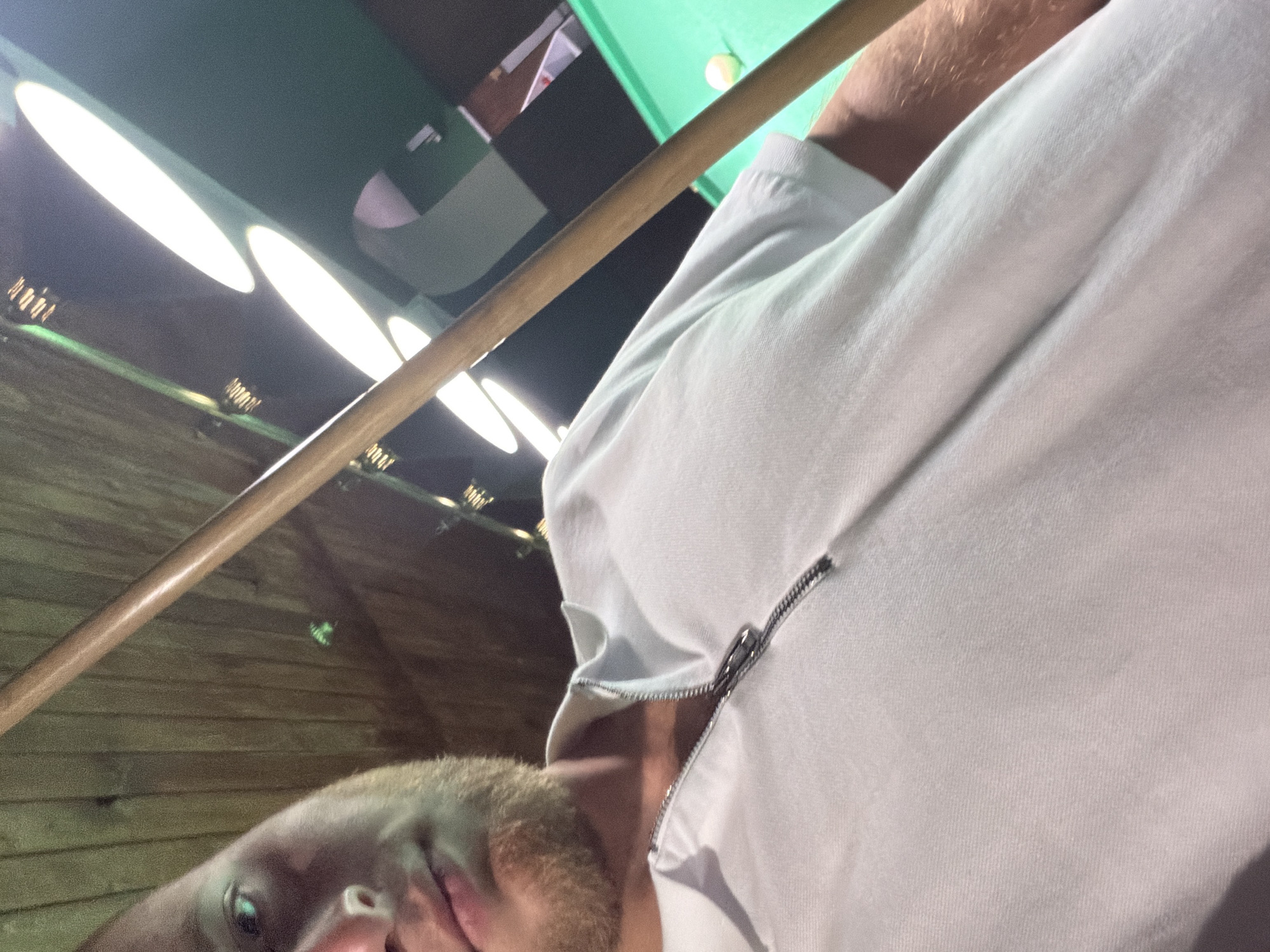 The image shows a close-up, first-person perspective of a man, likely in his early to mid-20s, with light skin and a short beard, holding a pool cue. He is wearing a white polo shirt with a zipper closure at the neck. The background suggests an indoor setting, possibly a billiard hall, with a green pool table visible, along with several large, oval-shaped overhead lights. The ceiling appears to be made of dark wood planks. The man's left eye, nose, and mouth are partially visible. The scene does not provide any specific indicators that would place it within the city of Strășeni, Moldova.