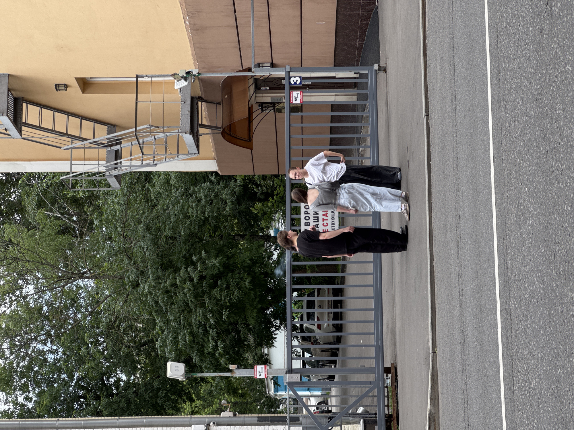 The image shows an outdoor scene in Moscow, Russia, with a metal gate and a building in the background. Three individuals are standing near the gate, engaged in conversation. The person on the left wears a black t-shirt and black pants, the person in the middle wears a grey long-sleeved shirt and a light blue skirt, and the person on the right wears a white t-shirt and black pants. Behind them, cars are parked. The building has a light yellow facade and a metal fire escape. A sign on the gate displays the number "3" and a red square with a camera icon. The environment appears to be an urban setting with trees and a sidewalk.