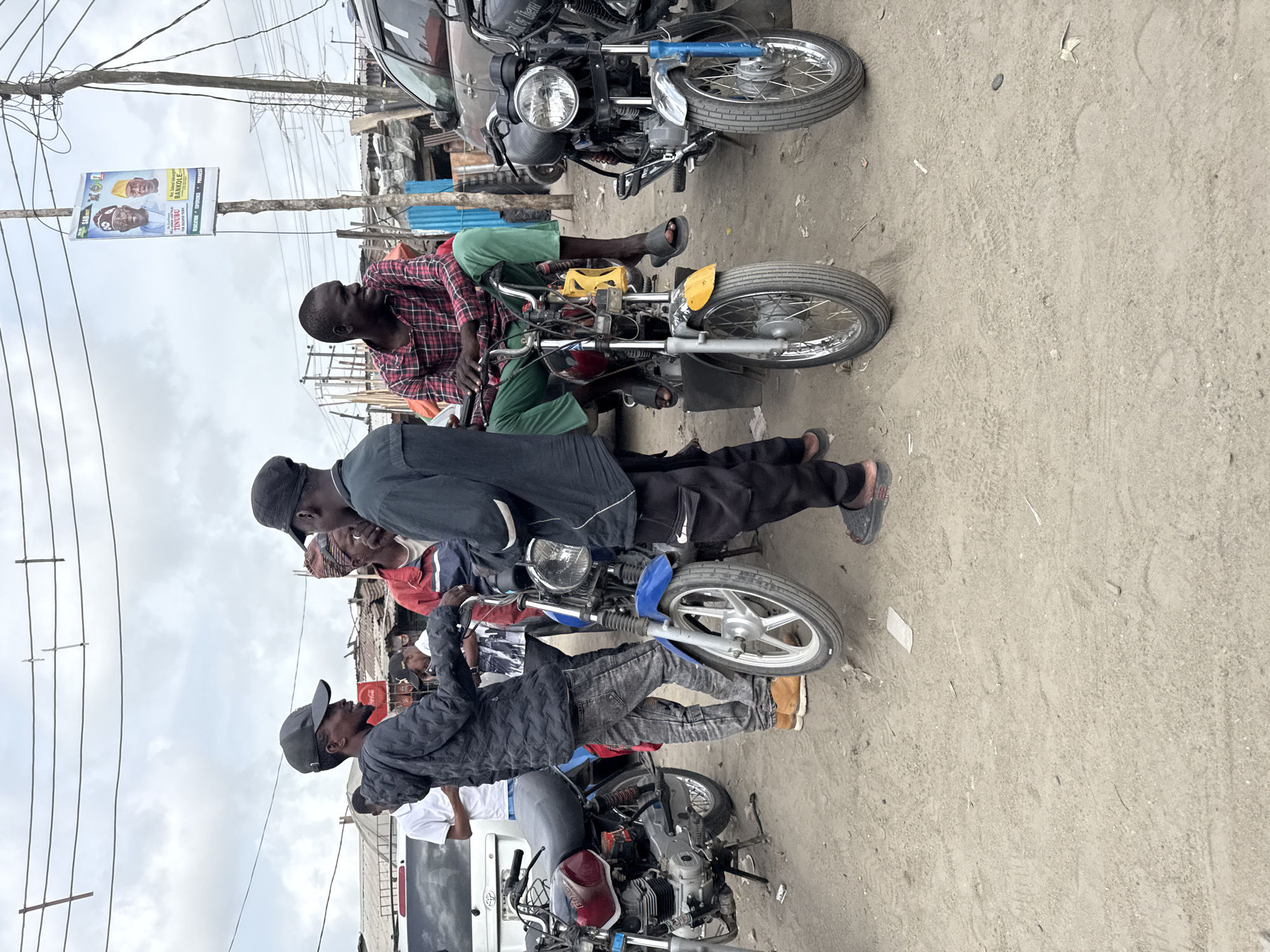 The image depicts a street scene in what appears to be a commercial area of Lagos, Nigeria. Several motorcycles are parked or stopped on a dusty, unpaved surface. A group of men are gathered around the motorcycles, some are sitting on them. The men appear to be of African descent, wearing casual clothing. A political campaign poster for Tinubu and Bankole is attached to a utility pole in the background. The background also shows cluttered storefronts, power lines, and a cloudy sky.
