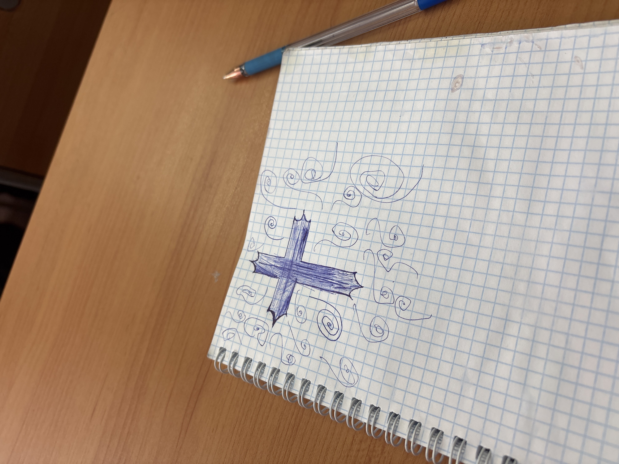 The image displays an angled, slightly elevated view of a light brown wooden desk surface, upon which a spiral-bound notebook and a blue ballpoint pen are situated. The desk's surface features a distinct wood grain pattern.
A white spiral-bound notebook with pages featuring a light blue grid pattern (squared paper) occupies the right half of the frame. Its white plastic or metallic spiral binding is visible along its left edge. Resting diagonally across the top left corner of the notebook, and partially extending onto the desk, is a blue ballpoint pen with a clear barrel.
The notebook page prominently displays a doodle in blue ink. The central element of this drawing is a stylized, thick blue cross, entirely filled with pen strokes. The four arms of the cross terminate in sharp, pointed extensions, giving it a distinctive, almost serrated look. Surrounding this central cross are numerous smaller, freehand doodles, consisting mainly of various swirling spirals and undulating wavy lines, all drawn in the same blue ink. There are minor smudges or discolorations along the top edge of the notebook page.
No people are visible in the image. The setting appears to be an indoor environment, likely a study, classroom, or office desk, during what seems to be daylight hours or under bright artificial lighting. No discernible text is present on any of the objects.