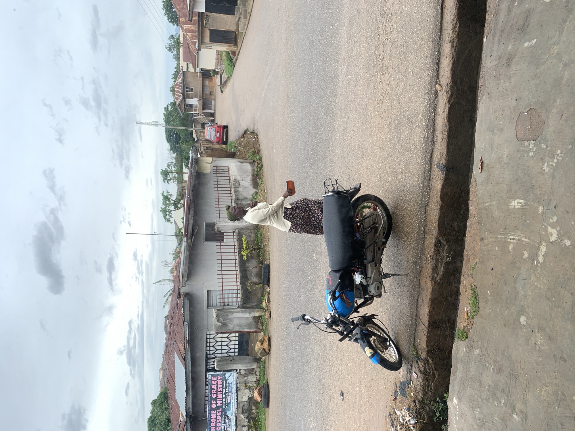 This vertical-format image presents an outdoor street scene, likely in a residential or commercial area of Jos, Nigeria. The main subjects are an elderly woman and a parked motorcycle.
The woman stands beside a blue and black motorcycle, which features a large black carrier or storage box on its rear. She is wearing a light-colored, long-sleeved shirt, a dark patterned skirt, and a head covering. In her right hand, she holds a small, rectangular brown object, possibly a container or wallet.
The paved road appears worn in places, with a concrete curb and an open drainage ditch running alongside it. Several modest buildings, some with corrugated metal roofs and security grilles, line the street. A red car is visible further down the road. The sky is overcast with gray clouds, indicating a cloudy day with soft, diffused lighting.
On a building to the left, a sign is clearly visible with the text: "THRONE OF GRACE GOSPEL MINISTRY," "HIS PRESENCE SCHOOL OF THE SWORD," and "(Infinity Bible Class)." The overall atmosphere suggests a typical street scene in a local community.