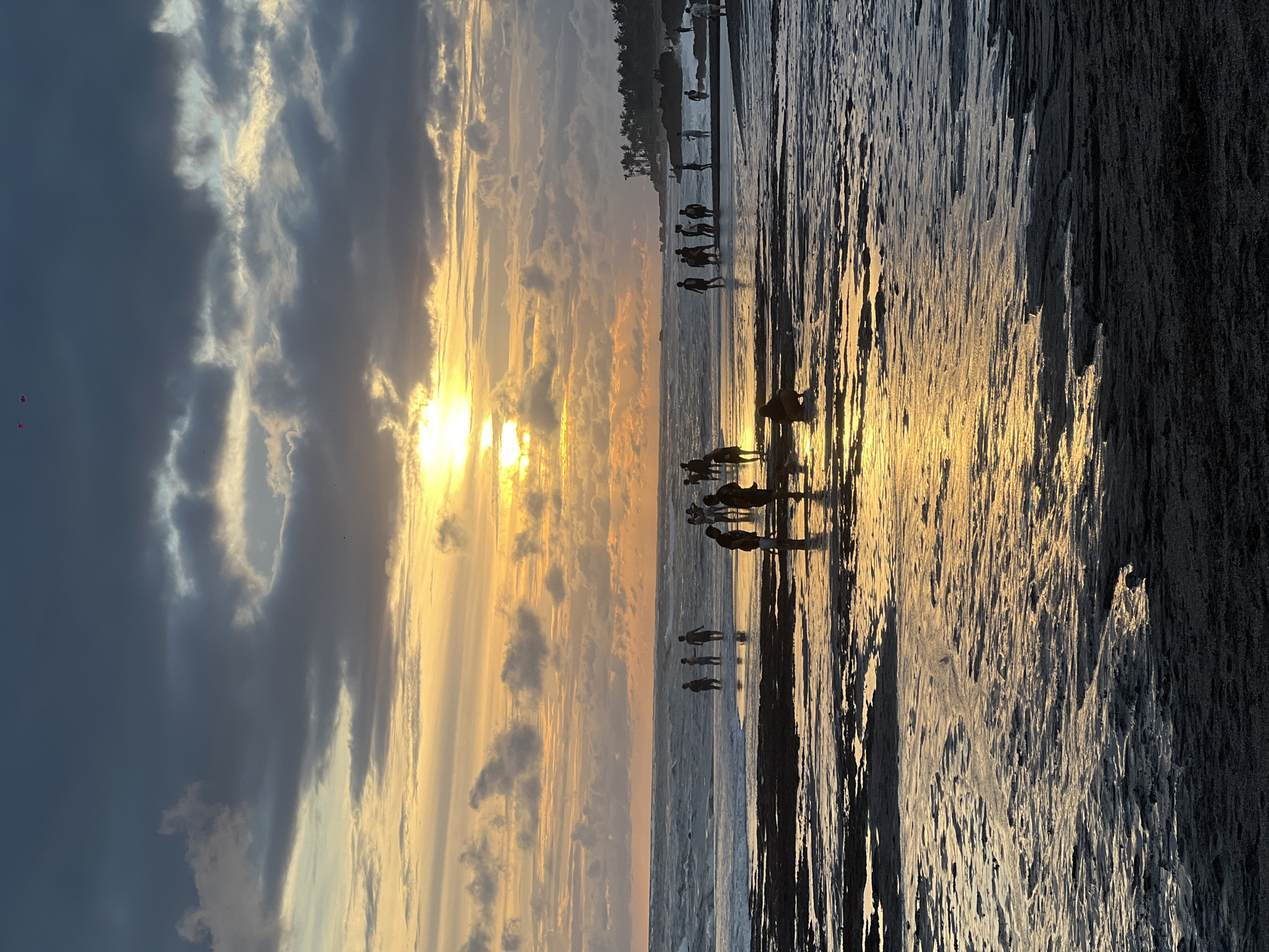 The image depicts a sunset scene at Mengwi, Indonesia. Silhouetted figures of people are visible along the shoreline and in the shallow water. The sun is setting behind a layer of clouds, casting a golden light that reflects on the wet sand and water. The sky is a mix of grey and golden hues due to the clouds and sunset. The ground is dark, likely wet sand, and reflects the golden light.