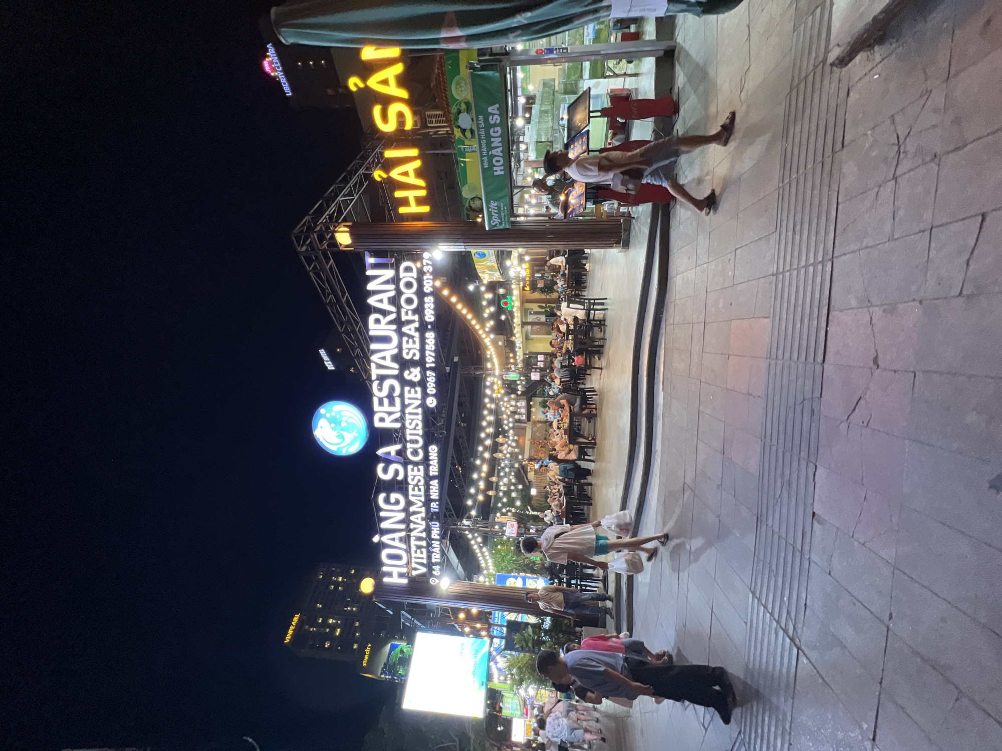 The image shows a nighttime scene in Nha Trang, Vietnam, specifically in front of "HOÀNG SA RESTAURANT VIETNAMESE CUISINE & SEAFOOD" located at 64 Trần Phú, TP. Nha Trang. The restaurant features a prominent illuminated sign and is decorated with numerous string lights. People are seen walking on the sidewalk and seated at tables within the restaurant, suggesting a busy dining establishment. Several buildings are visible in the background, including "VINPEARL" and "LIBERTY CENTRA", indicating an urban setting. The ground in the foreground is paved with large stone tiles and has a tactile paving strip. A man in a white t-shirt and shorts, carrying two plastic bags, is walking towards the right side of the frame.