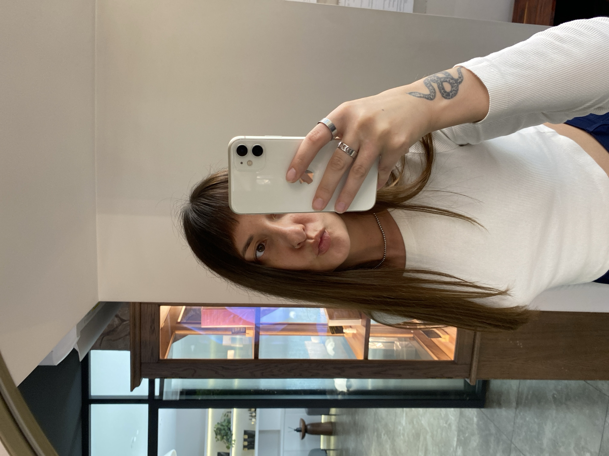 In Votkinsk, Russia, a young woman with long, brown hair and bangs takes a mirror selfie. She is wearing a white, long-sleeved, ribbed top and dark blue bottoms. The woman has a nose ring and is wearing two rings on her right hand, one on her index finger and one on her middle finger. A tattoo of a snake is visible on her left forearm. She is holding a white smartphone to take the photo. The background shows a reflection of a room with a wooden display cabinet filled with items, and a window that looks out onto an indoor space with shelves and seating. The lighting is bright and appears to be artificial.