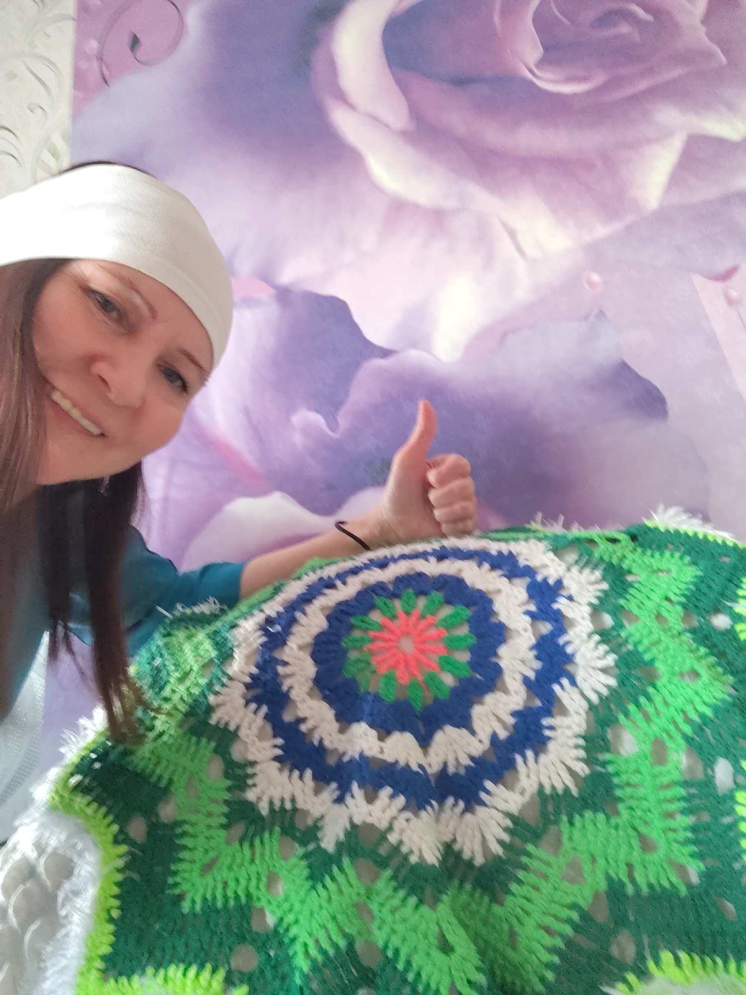 A smiling woman is partially visible in a close-up indoor shot, proudly presenting a colorful, circular crocheted item. She has long dark hair and wears a white headband and a teal or blue long-sleeved top. Her face, seen from the lower-left, beams with a wide smile, and her right hand gives a clear thumbs-up gesture, indicating approval or satisfaction.
The crocheted piece features a vibrant pattern, starting with a pink or orange star-like center, encircled by concentric layers of green, blue, and white yarn, predominantly in various shades of green on the outer edges. This handmade item, possibly a blanket or doily, shows intricate crochet work with an open, textured design.
The background is a wall adorned with wallpaper displaying large, soft purple or lilac floral motifs, resembling roses or similar blossoms. A lighter, subtly textured wall is visible on the far left of the frame. The overall scene suggests a domestic setting where the woman is showcasing her creative work. Her smile and the thumbs-up gesture convey happiness, satisfaction, and possibly pride in her handicraft. No text is visible in the image. The implied location is a domestic setting in Сусады-Эбалак, Russia.