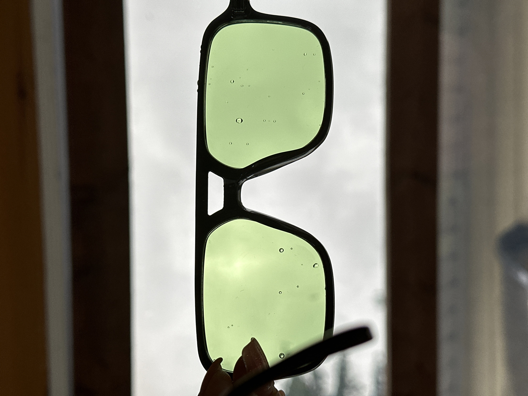 The image shows a pair of black-framed sunglasses with light green lenses being held up against a window. There are several small bubbles visible within the lenses. The background seen through the lenses is a blurry, overcast sky. Parts of a window frame and possibly a curtain are visible on either side of the sunglasses. A finger is visible holding the glasses. There is no way to identify the location as Nizhny Novgorod, Russia from the content of the image.