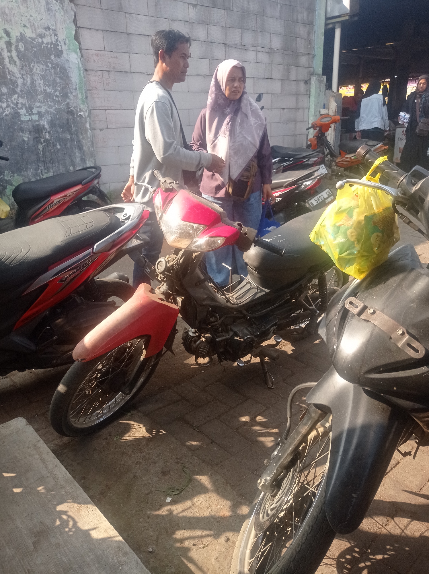 Two individuals, a male and a female, stand between several parked motorcycles on a paved ground surface. The male, on the left, wears a light grey long-sleeved shirt and dark pants, with a bag strap visible across his chest. The female, to the right of the male, is wearing a light purple hijab and a dark purple top, with a small brown bag slung across her shoulder. She appears to be looking towards the male.
Several motorcycles are present. In the foreground, a red and black motorcycle is prominently displayed, its front wheel and fender visible. To its right, the front of a black motorcycle is visible. Behind these, more motorcycles can be seen, including a red one with black accents and an orange one. A bright yellow plastic bag hangs from the handlebars of one of the motorcycles.
The background features a textured wall constructed of grey rectangular blocks on the left, with some green moss or paint patches. To the right, there is an overhead structure with supporting columns, suggesting a covered area, possibly a market or roadside stall. The scene appears to be outdoors, with sunlight casting distinct shadows on the ground. The license plate on one of the motorcycles reads "N 5892 CR." The overall impression is that of a public area, likely a street or a market in Tajinan, Indonesia, where people are gathered around parked vehicles.