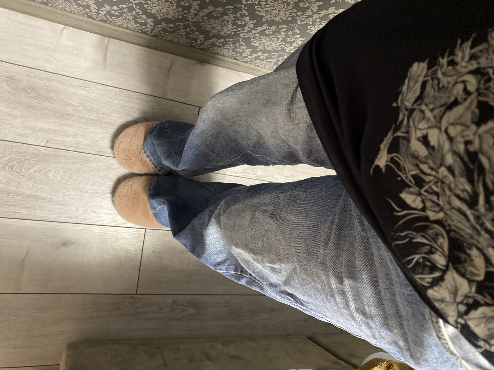 The image shows a person's legs from an overhead perspective. The person is wearing light blue jeans and tan slippers. The person is also wearing a black garment with a white floral pattern. The background consists of a light-colored wooden floor and wallpaper with a floral design. There is no information in the image to confirm or deny that the image was taken in the city of Ternopil, Ukraine.