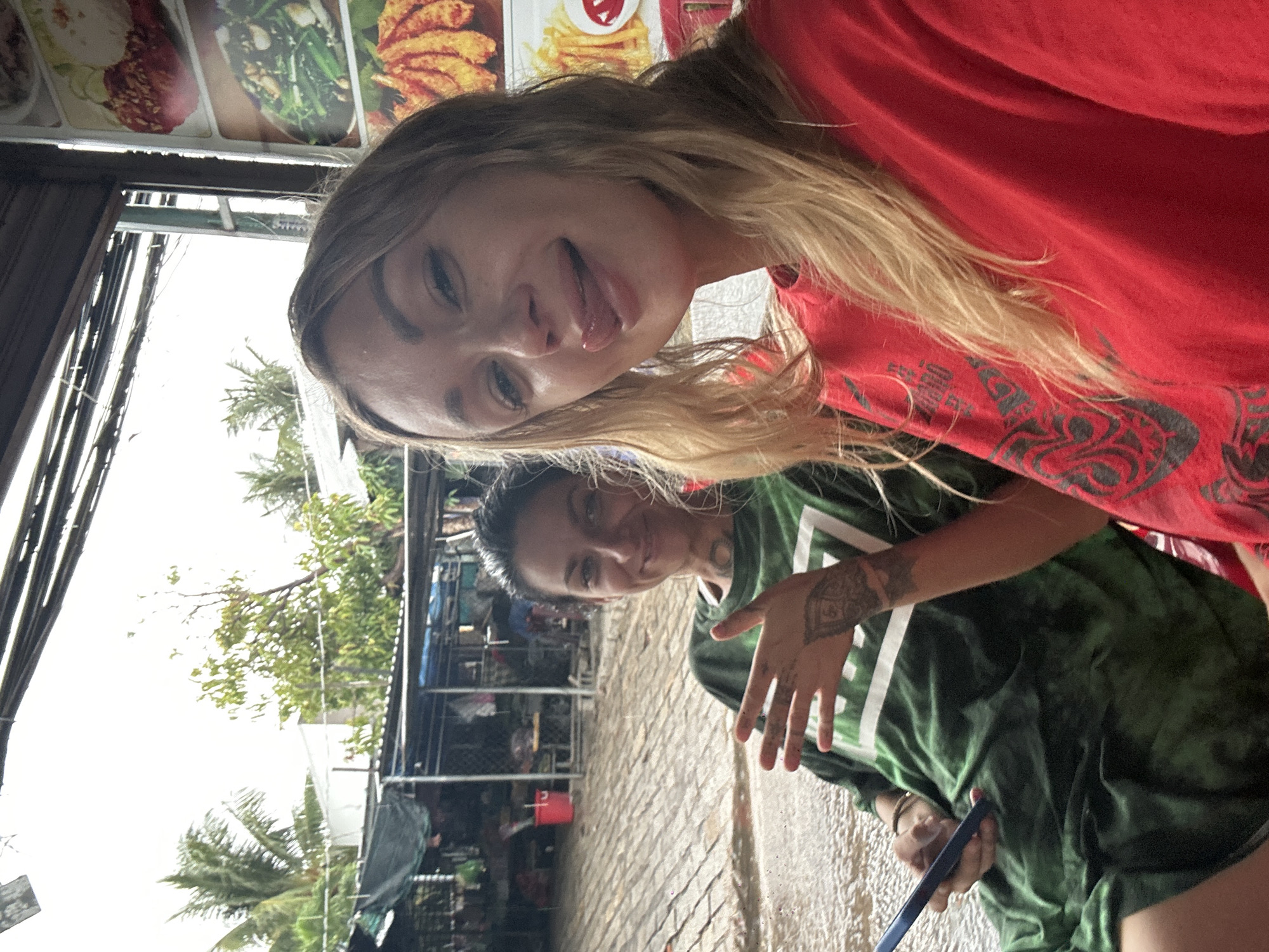 Two females are photographed in what appears to be Phu Thuy Ward, Vietnam. One wears a red shirt with a decorative print. The other wears a green shirt. Both face the camera. The setting includes a paved street and storefronts, with foliage visible in the background.