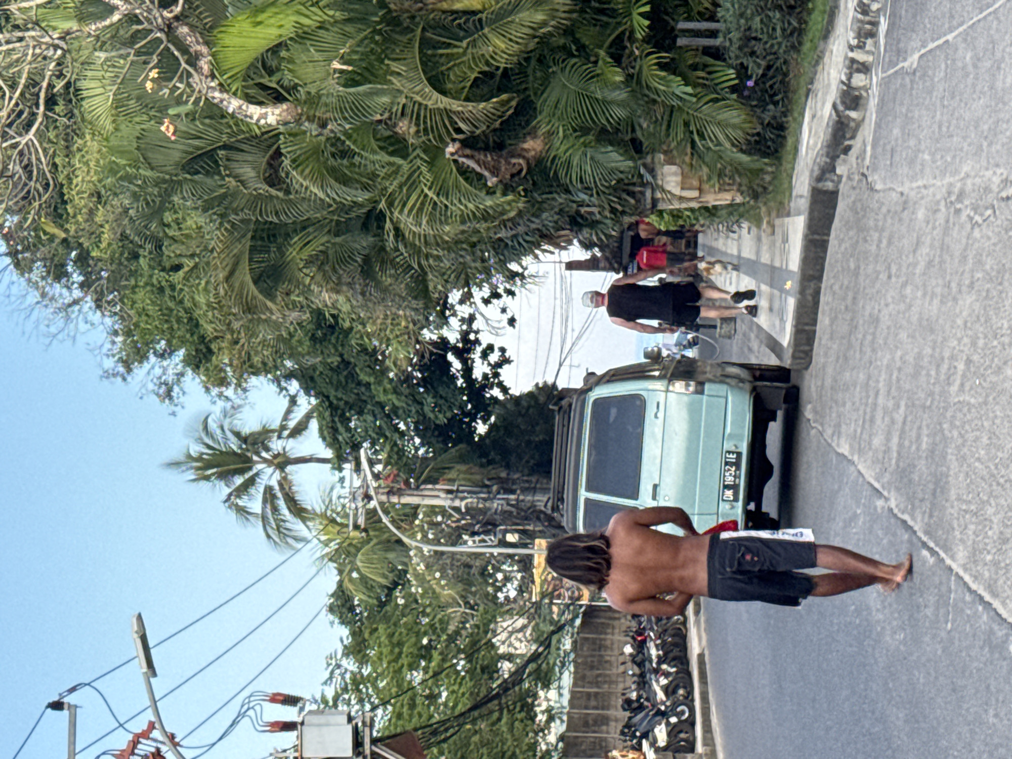 This image captures a street scene in Kuta, Indonesia, characterized by tropical vegetation and urban infrastructure. The foreground features a man with long, dark hair, shirtless and wearing dark shorts with a white panel, walking barefoot on the asphalt road away from the viewer. He holds a white object in his left hand. To his right, a light blue minivan with a roof rack is parked, displaying the Indonesian license plate "DK 1952 IE."
Further along the road on the right, a man wearing a white cap, dark sleeveless shirt, and dark shorts walks on a concrete sidewalk, also facing away from the viewer. He is walking a small, light-colored dog on a leash. Behind him, another individual in a red shirt is partially visible, walking further down the sidewalk. The sidewalk on the right is bordered by dense tropical foliage, including prominent palm trees and other green plants, which line the street and create a lush backdrop.
On the left side of the street, numerous motorcycles of various dark colors are parked parallel to the road, extending into the mid-ground. Overhead, multiple utility poles support a complex network of black power lines and a grey street lamp. A large, grey utility box is mounted on one of these poles. The sky above is clear and light blue. The overall scene depicts a busy street environment with a mix of pedestrian activity, parked vehicles, and local infrastructure under a bright sky.