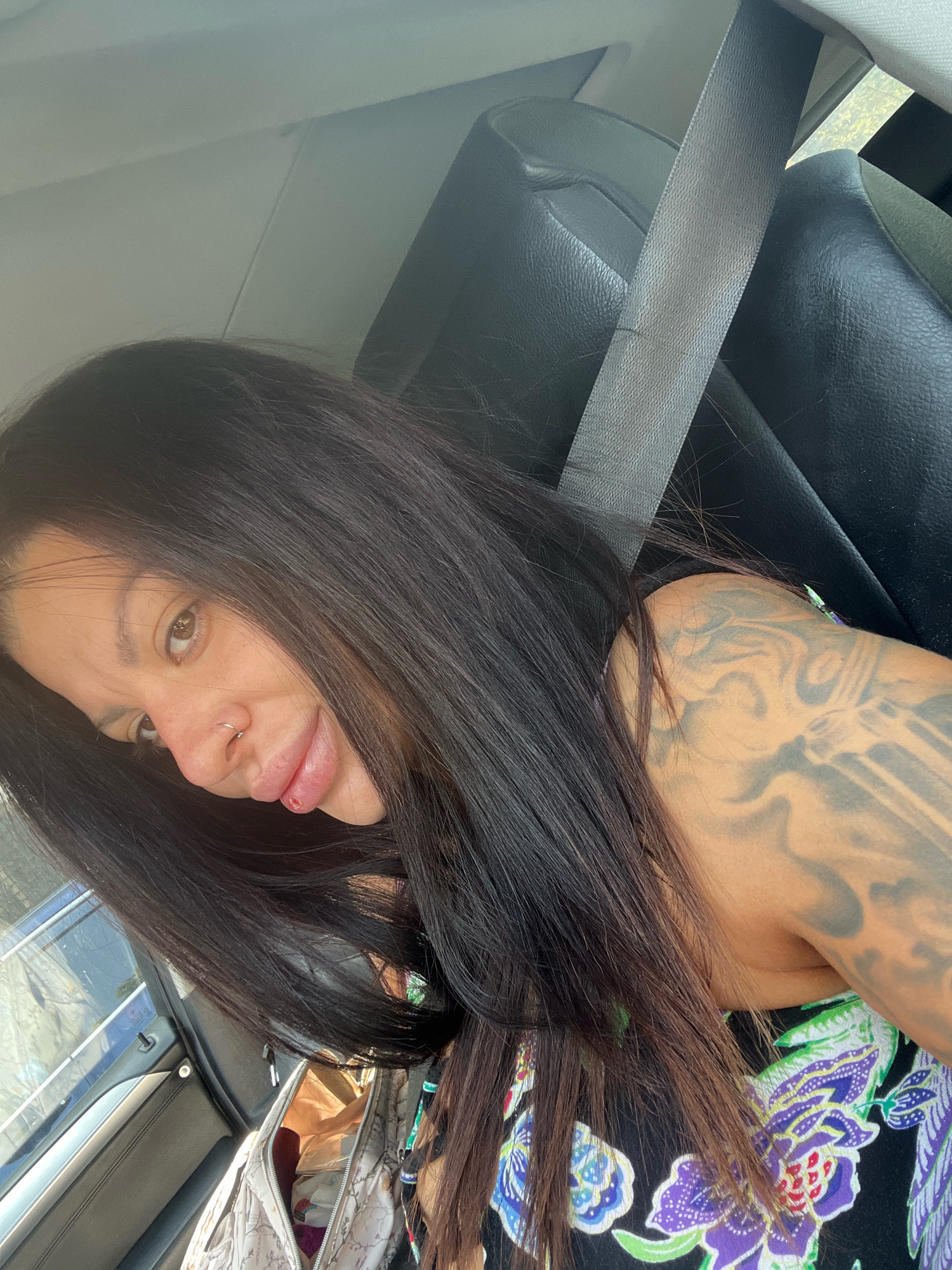 A woman with long dark hair and tattoos on her arm is sitting in the passenger seat of a car. She is wearing a floral print dress. The car appears to be in motion. The background is blurred, suggesting the photo was taken while the car was driving. The image does not provide any information that could be used to determine the location in the city of Plovdiv, Bulgaria.