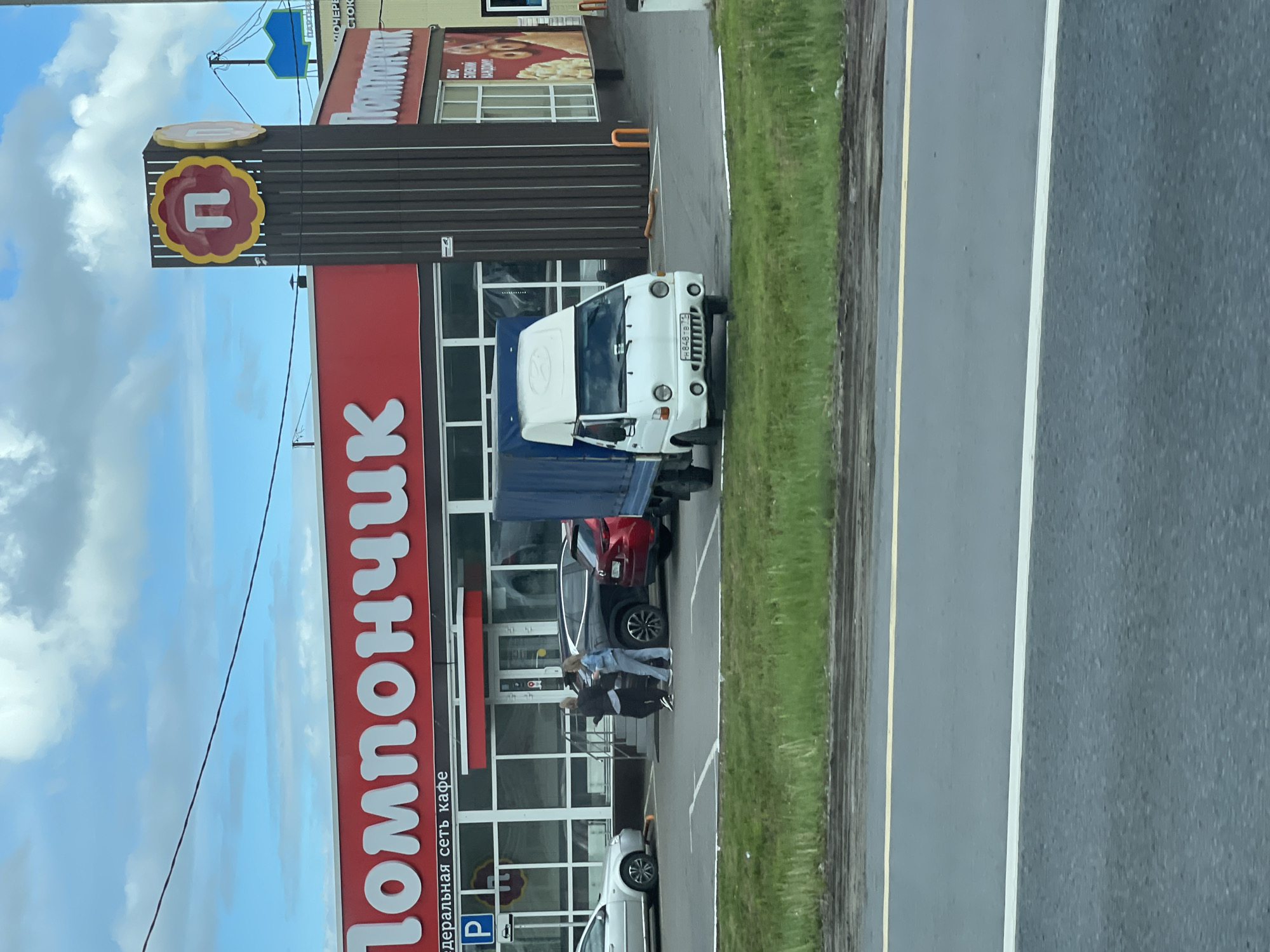 A vertically oriented medium shot captures a "Pomponchik" (ПOMПOHЧИK) cafe in Berezhki, Russia. The building is red and white, with windows. A red and yellow logo is on the building. A white truck is parked outside near the street. There are a couple of people standing near the entrance and vehicles parked in the parking lot. The sky has blue and white clouds. The street is gray. The grass is green.