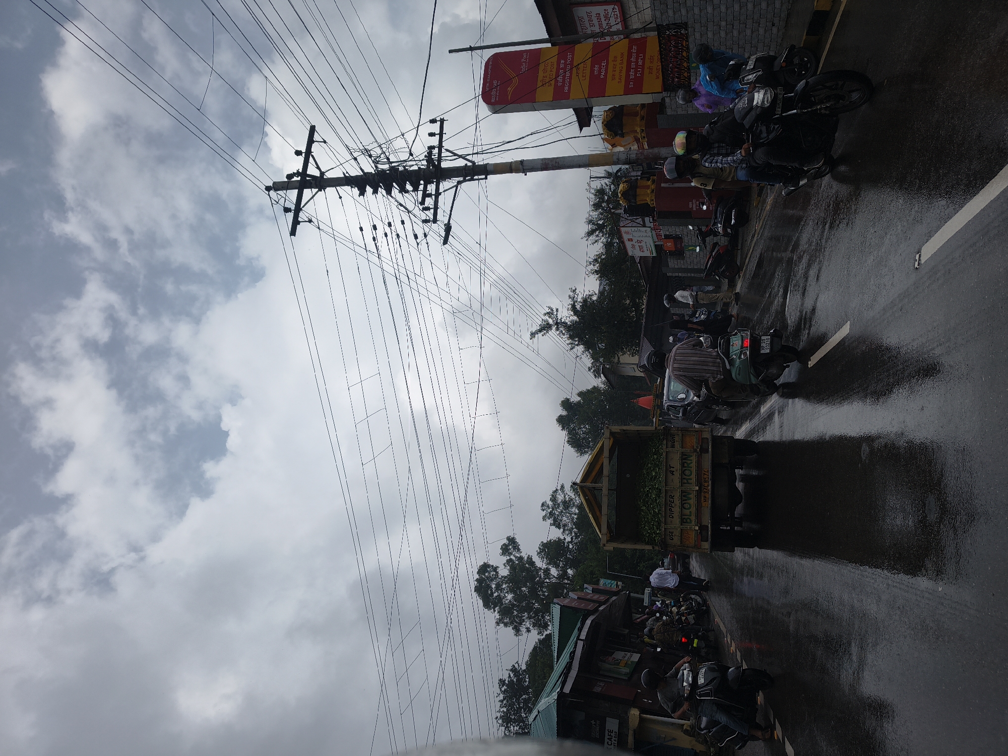 The image depicts a street scene in Dharamshala, India, under an overcast, cloudy sky, suggesting potential rain or recent rainfall as the road surface appears wet and reflective. The environment is a commercial street with buildings lining one side, featuring signs in Hindi. A prominent utility pole with numerous electrical wires is visible on the right side of the frame, along with a large red and yellow sign for "India Post" and a "Saving Bank."
Several vehicles are present, including a truck filled with green foliage (likely tea leaves), multiple motorcycles, and what appears to be a scooter. People are seen riding or standing near these vehicles. On the left, a person is riding a black scooter. In the middle, a truck is stopped, and to its right, several motorcycles are parked or moving. Further to the right, a black motorcycle with two riders passes by, and a person in a blue jacket is leaning over another motorcycle. A few other individuals are walking or standing on the side of the road. The overall impression is of a busy urban or semi-urban street during a potentially damp day.