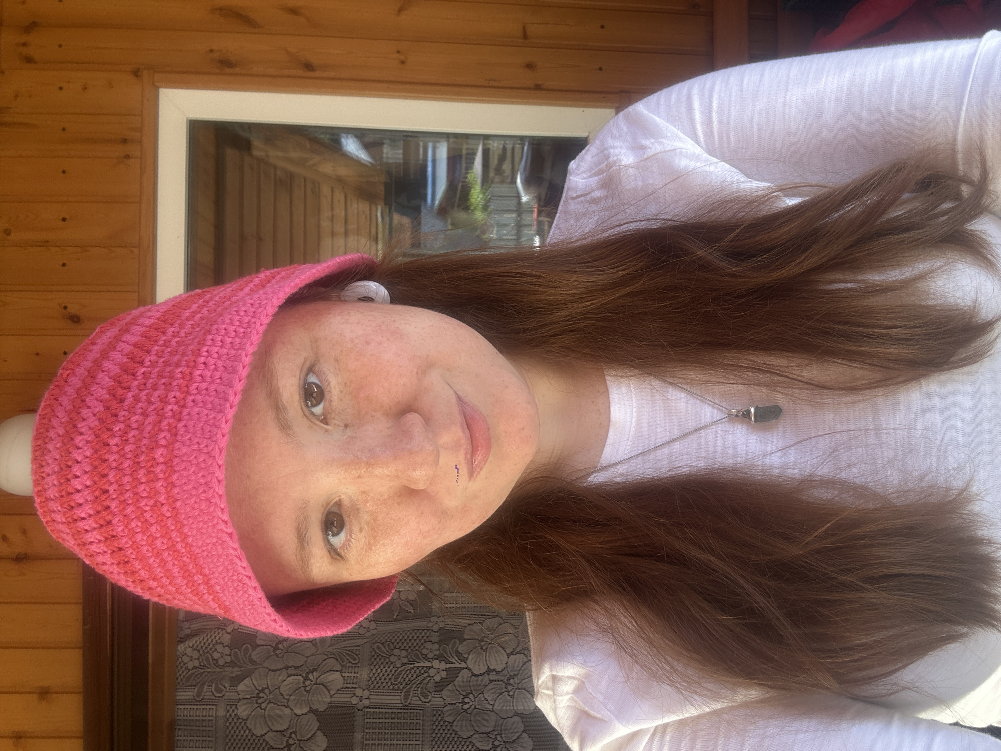 A close-up, chest-up portrait features a person with fair skin and numerous freckles, gazing directly at the camera with a gentle smile. They have long, wavy reddish-brown hair that extends past their shoulders. A vibrant pink knitted beanie covers their head. The person is dressed in a white long-sleeved top, adorned with a silver chain necklace holding a dark, possibly stone, pendant. Small white earbuds are visible in their ears, and a subtle stud piercing is present on their left nostril.
The background primarily consists of light brown wooden paneling, suggesting an outdoor structure like a cabin, patio, or porch. A white-framed window is visible, reflecting greenery and what appears to be part of a building, indicating a bright, outdoor setting. Natural, bright lighting illuminates the scene, consistent with a sunny daytime. A dark fabric with a floral or lace pattern is partially visible in the lower left portion of the image. The overall expression conveys a calm and pleasant demeanor. The location is Nizhny Novgorod, Russia. There is no visible text.