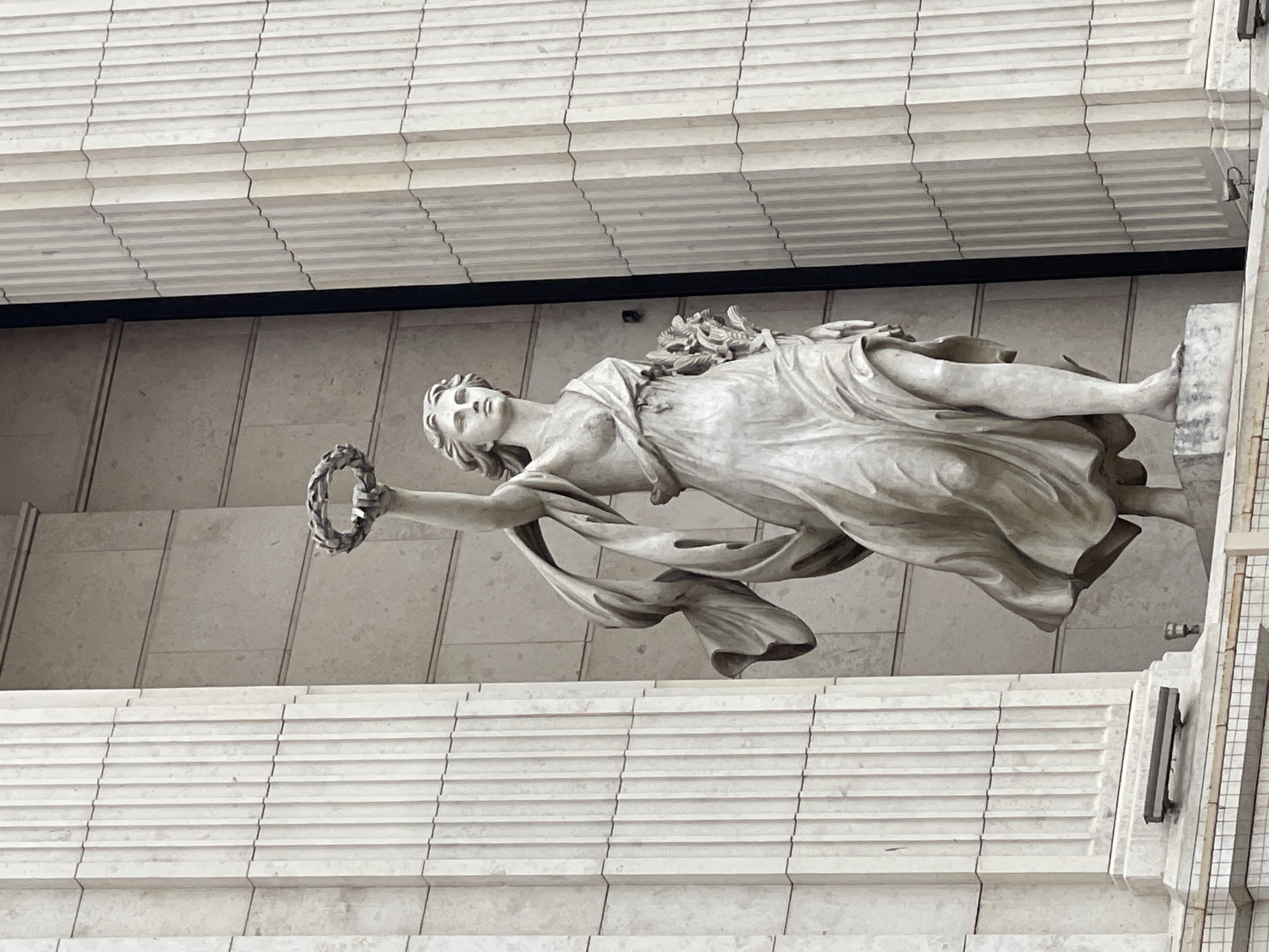 The image shows a statue of a female figure, possibly a goddess or allegorical figure, in Saint Petersburg, Russia. She is draped in flowing robes and holding a laurel wreath in her outstretched arm. The statue is mounted on a building facade, with horizontal architectural details visible in the background. The statue is made of stone and appears to be part of a larger architectural composition.
