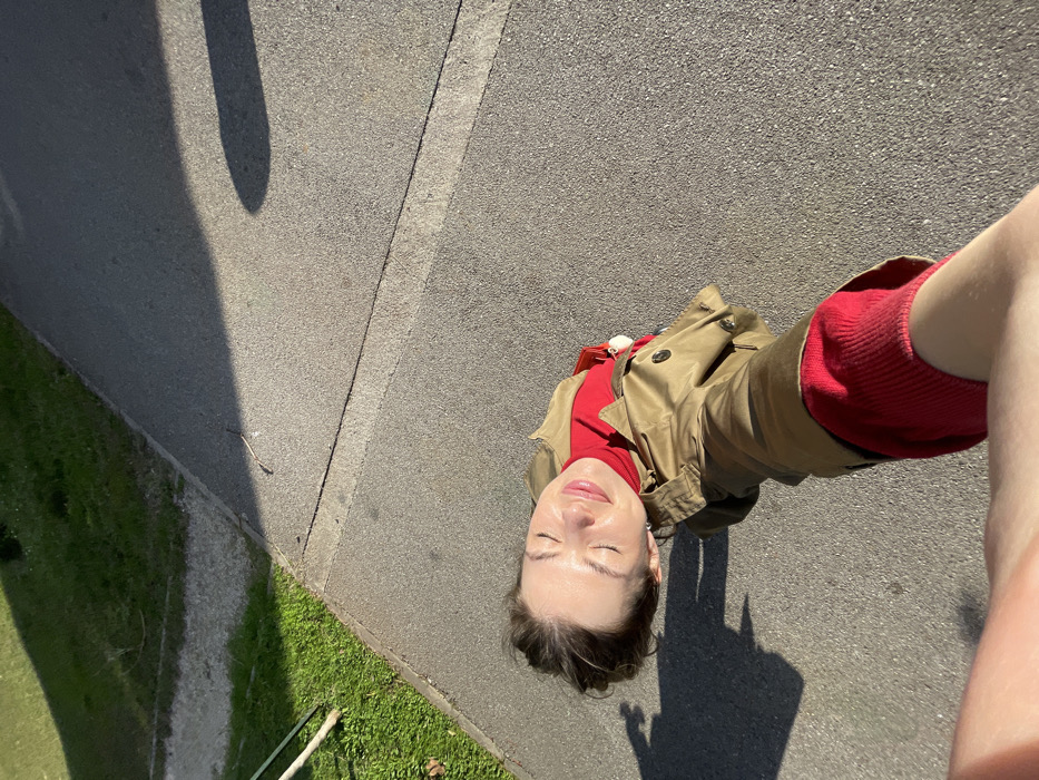 A high-angle selfie depicts a female subject outdoors in Budva, Montenegro, under strong sunlight. The subject, with light complexion and eyes closed against the sun, wears a red top and an olive-tan trench coat, holding the camera with her right arm extended into the lower left frame. She is positioned on a light grey asphalt pathway, which borders a strip of green grass on the left. A slender stick rests at the junction of the pavement and grass. Prominent shadows are cast by the subject's head on the pavement to her left, and a large, elongated shadow occupies the upper right portion of the paved area and grass.