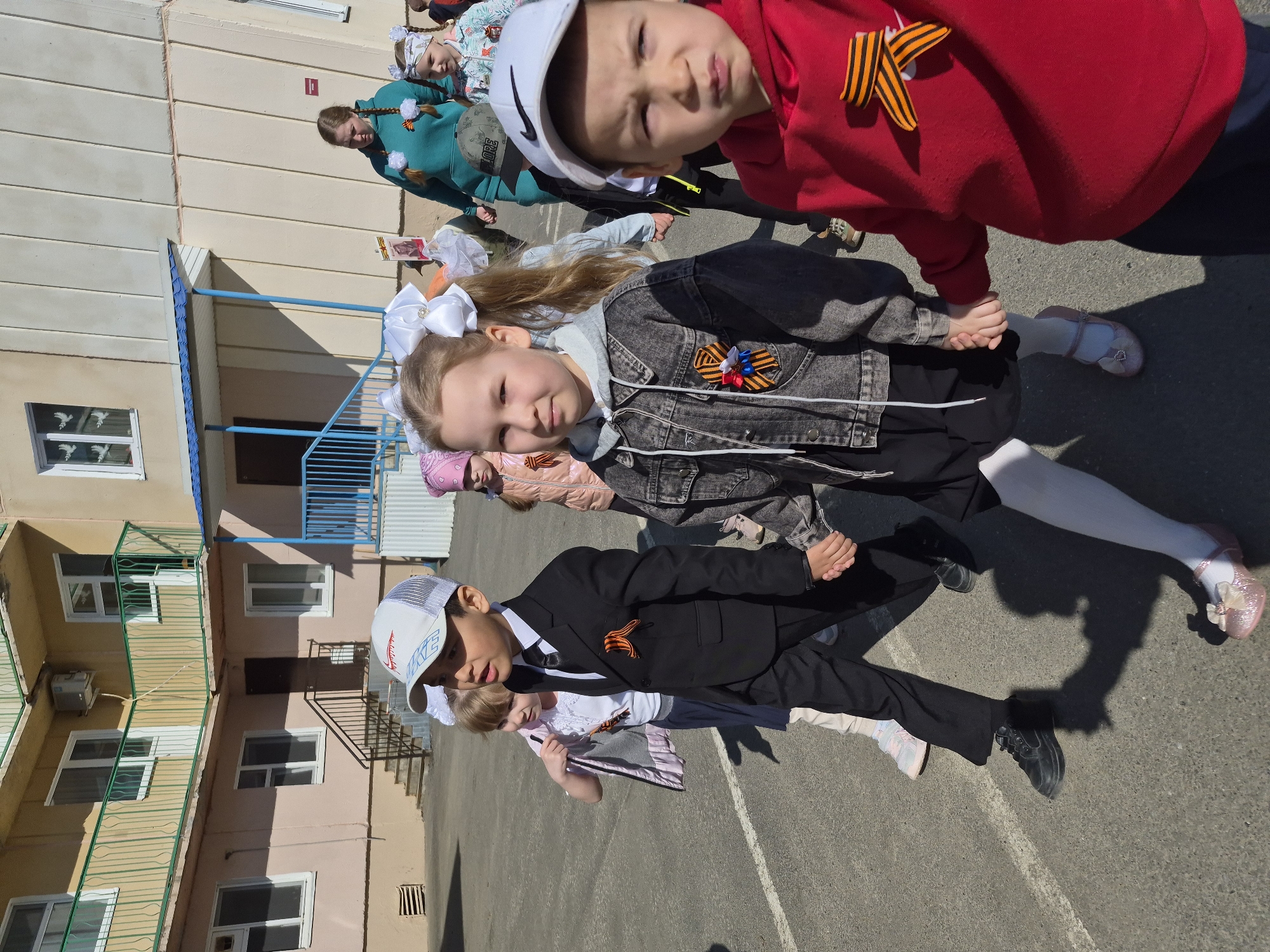 A group of children, possibly students, are walking in an outdoor area next to a multi-story building. The building is light pink and yellow, with multiple windows and metal railings. At least four children are clearly visible:
* A boy wearing a black suit, white shirt, black shoes, and a baseball cap. He has an orange-and-black ribbon pinned to his jacket.
* A girl with white bows in her hair, wearing a black denim jacket over a black dress and white tights. She is holding hands with a boy.
* A boy wearing a red hooded sweatshirt and a white baseball cap with an orange-and-black ribbon pinned to his shirt.
* Other children are partially visible in the background.
The children are walking on a paved surface with white lines. The atmosphere appears bright and sunny.
The context of the image suggests this is likely a Victory Day celebration.
It is not possible to ascertain the precise location in Tobolsk, Russia, solely from the visual information in the image.