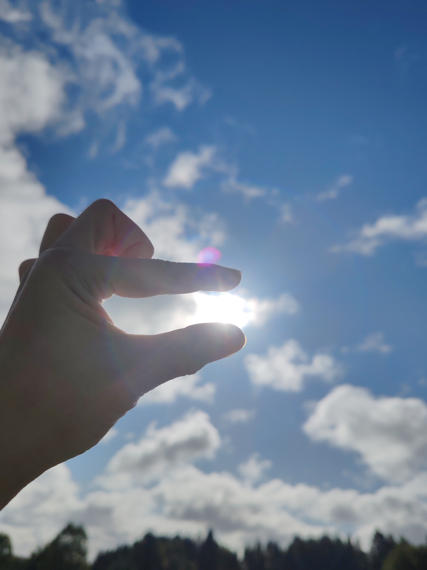 A hand is silhouetted against a bright sky, with the sun shining through the gap between the thumb and forefinger. The sky is blue with fluffy white clouds, and a faint lens flare is visible. In the background, a dark line of trees suggests a distant horizon, possibly indicating a location like the city of Chuhloma, Russia, where such a landscape might be found. The focus is on the hand and the sun, with the background slightly blurred.