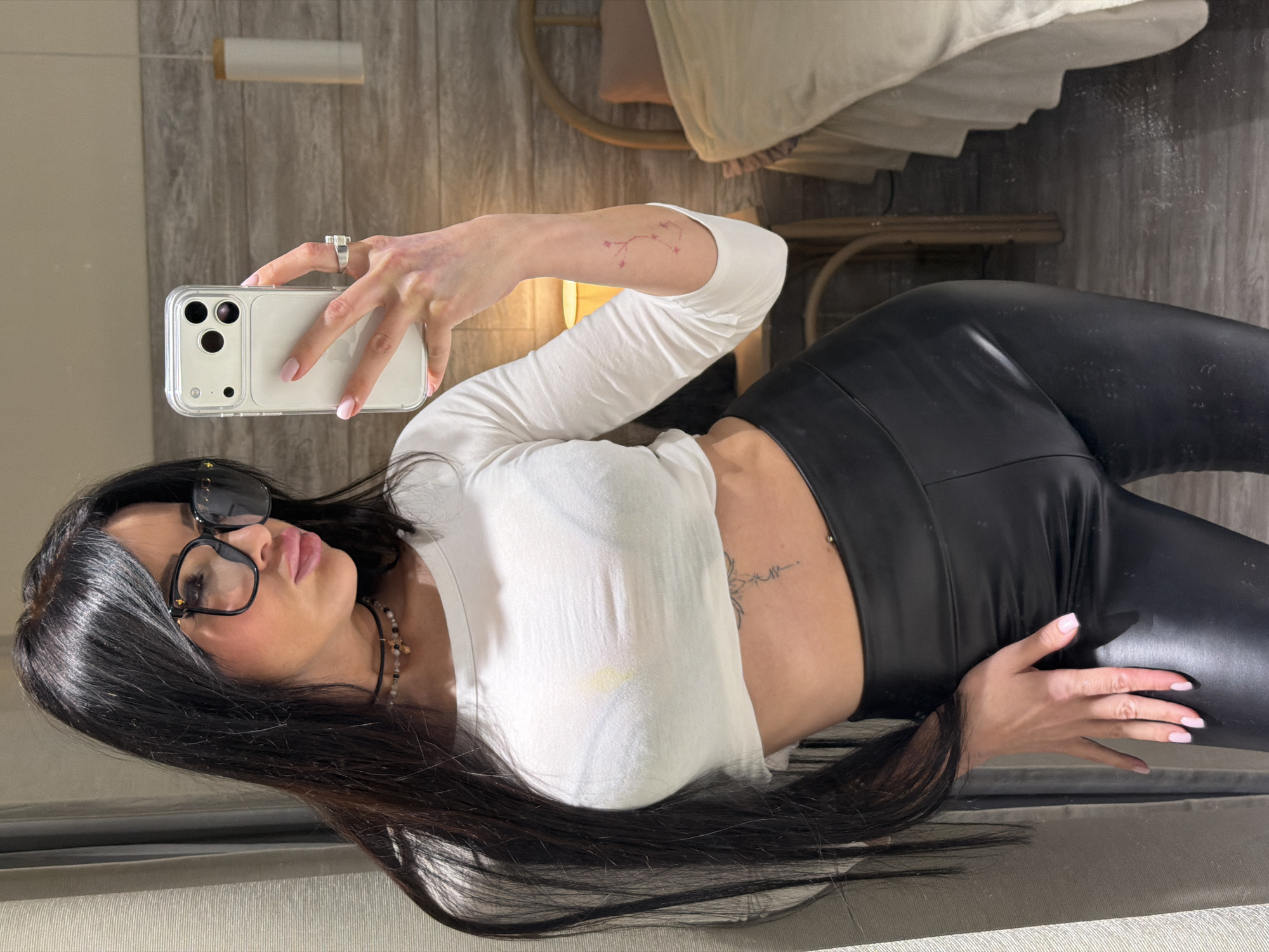 In an indoor setting in Moscow, Russia, a woman takes a mirror selfie. The image captures her from the chest up to her lower thighs, with the entire scene rotated 90 degrees counter-clockwise. She holds a light-colored iPhone, encased in a clear protective cover, in her right hand, which also displays a silver-toned ring on her ring finger and light pink nail polish.
The woman has long, dark hair and is wearing a white long-sleeved crop top that exposes her midriff, revealing a tattoo on her left side, possibly a floral or mandala design, and a belly button piercing. Another tattoo, resembling a constellation, is visible on her right upper arm. She is also wearing black, shiny faux leather leggings. Her face, framed by dark hair, is turned slightly to the side, showcasing full lips and black-framed glasses or sunglasses resting on her head. A multi-bead necklace is visible around her neck.
The background features a textured wall, possibly wood paneling, and part of what appears to be a bed frame or furniture made of light-colored wood or wicker. The lighting suggests an indoor environment. There is no discernible text visible in the image. The woman appears to be posing for the selfie, exhibiting a confident demeanor.