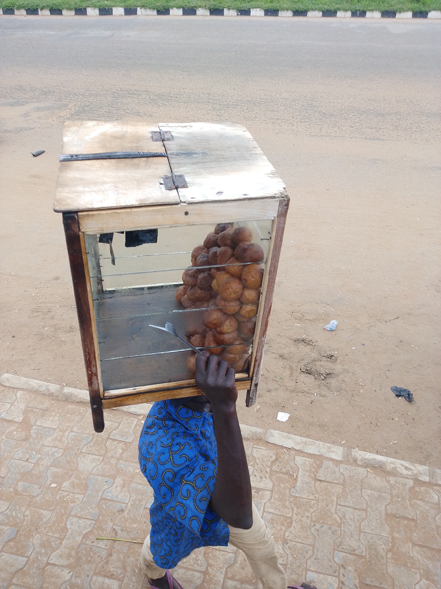 A person carries a wooden and glass display case filled with golden-brown fried dough balls. The case has hinges on top and is open at the front, revealing the pastry-filled interior. A hand reaches into the case, holding a fork. The person is wearing a blue garment with a yellow swirl pattern and light-colored trousers. The scene appears to be outdoors, with a dusty dirt ground and a paved sidewalk in the foreground. In the background, a road with distinct black and white stripes along its edge is visible, suggesting a roadside location. The lighting suggests it could be daytime, and the weather appears to be fair. The overall impression is of a street vendor, likely in a marketplace or along a busy route in Nigeria, given the location context provided.