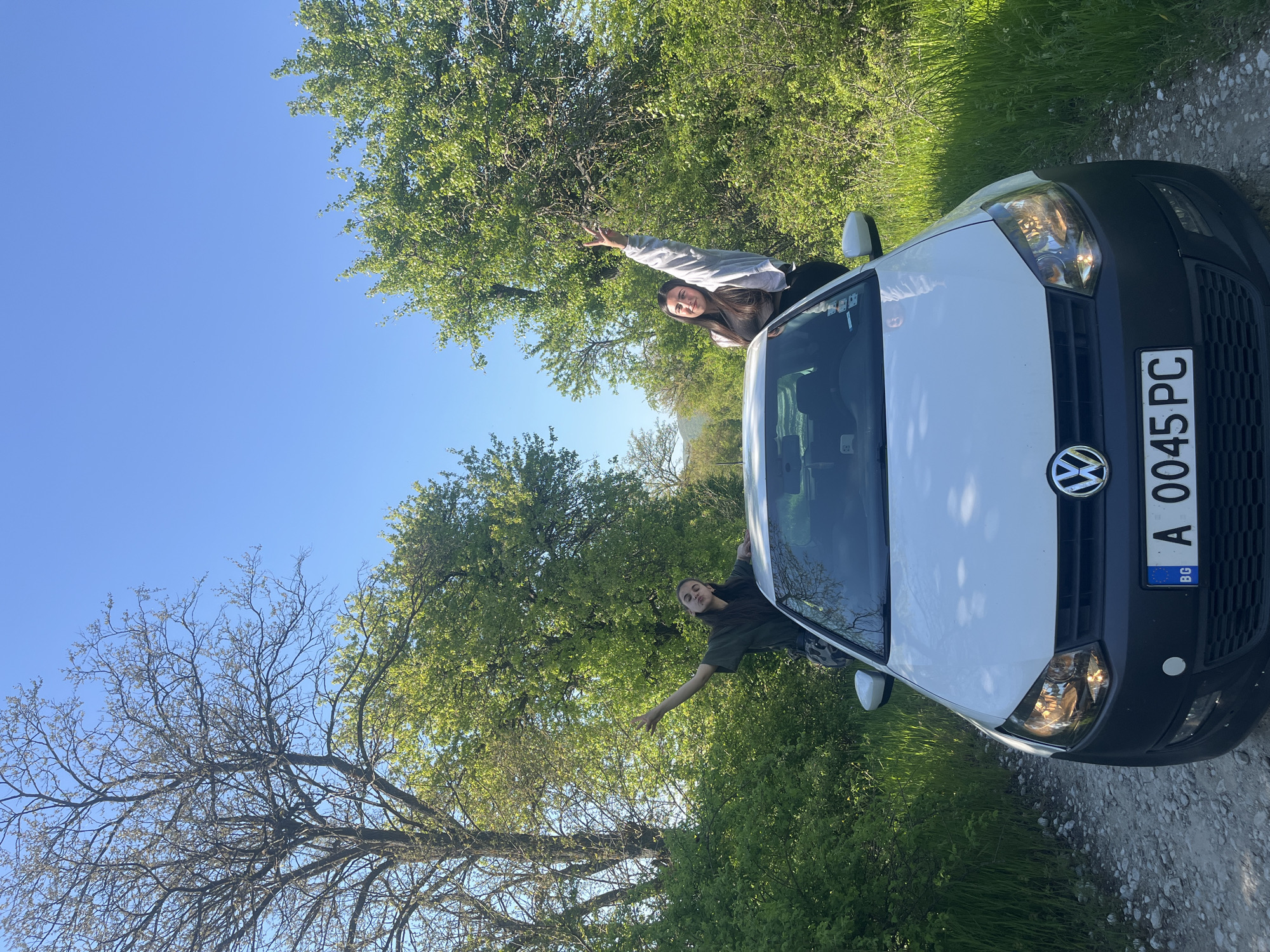 The image shows two people in a white Volkswagen car with the license plate "A 0045 PC" in Rish, Bulgaria. The car is parked on a dirt road surrounded by green trees and grass. The sky is blue. One person is leaning out of the driver's side window and the other out of the passenger side, both with their arms raised.