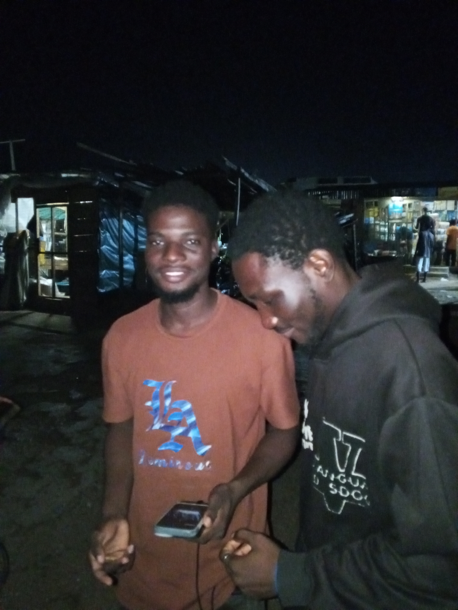 Two young men are standing in front of a row of informal businesses in Lokoja, Nigeria. The man on the left is smiling and wearing a reddish-brown t-shirt with a blue "LA" logo. He is holding a small, dark electronic device in his right hand. The man on the right is wearing a black hooded sweatshirt and is looking down, with his head tilted. The background shows the fronts of several small shops or stalls with some lighting. The scene appears to be outdoors at night.