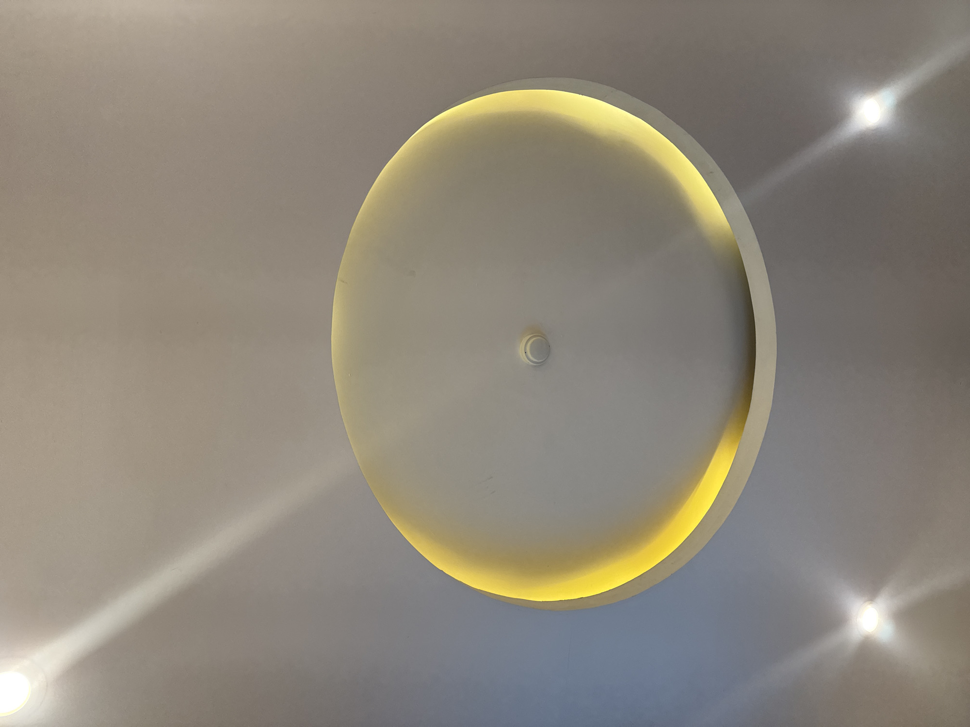 The image shows a ceiling with recessed lighting. In the center of the image is a circular recess in the ceiling, illuminated by a yellow light source. There are also two spotlights visible on the ceiling, one in the lower left and one in the lower right of the image. The ceiling is painted a neutral color, and the lighting creates a warm and inviting atmosphere. The image was taken in Hà Nội, Vietnam.
