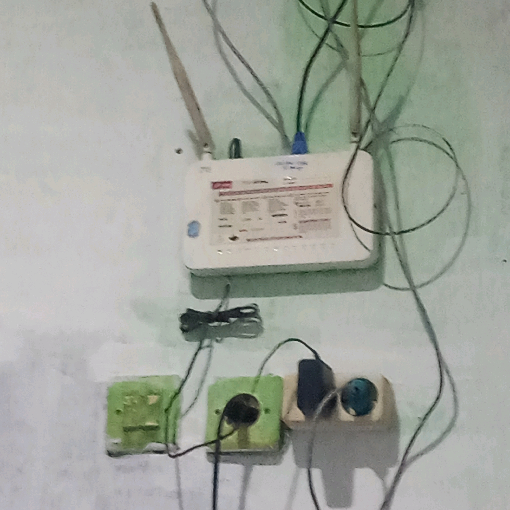 The image shows a close-up, overhead view of a router and electrical outlets mounted on a light-colored wall, likely in Kendayakan, Indonesia. The router, a white ZTE model, is centrally positioned at the top of the frame, with two antennas extending upwards. Various cables, including a blue Ethernet cable, are connected to the router and extend down the wall. Below the router, at the bottom of the frame, are three electrical outlets. The leftmost outlet is green and appears to be a standard outlet. The middle outlet is also green and has a black cable plugged into it. The rightmost outlet is beige and has a black power adapter plugged into it, with a blue indicator light visible. The overall scene is cluttered with wires and electrical devices. The wall appears to be textured and has a light green hue in some areas, possibly due to lighting or paint. The image is of moderate resolution and quality.