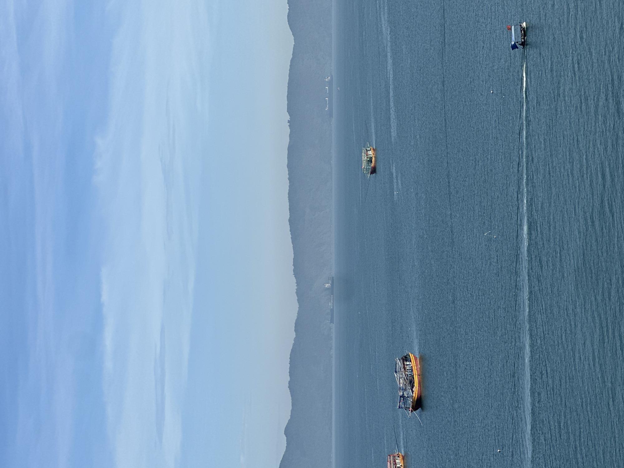 The image depicts a panoramic view of the sea, likely from a vantage point overlooking a coastline, characteristic of Đà Nẵng, Vietnam. The water is a deep blue, with gentle waves creating subtle ripples. In the distance, a chain of hazy, mountainous islands forms a backdrop against a pale blue sky with scattered, wispy clouds.
Several boats are scattered across the water. In the mid-ground, a large cargo ship, identifiable by the name "EVERGREEN" on its hull, is visible, suggesting active shipping routes. Closer to the shore, there are smaller vessels. One boat, with a yellow and red hull and a covered deck, is moored. Another, similar in design, is also present. Further out, a smaller boat is moving, leaving a white wake behind it. This boat appears to be carrying passengers and has flags displayed.
The overall scene conveys a sense of calm and vastness, with human activity represented by the boats navigating the expansive seascape. The clear, bright atmosphere indicates daytime, possibly morning or late afternoon, given the soft light and the haziness of the distant landforms.