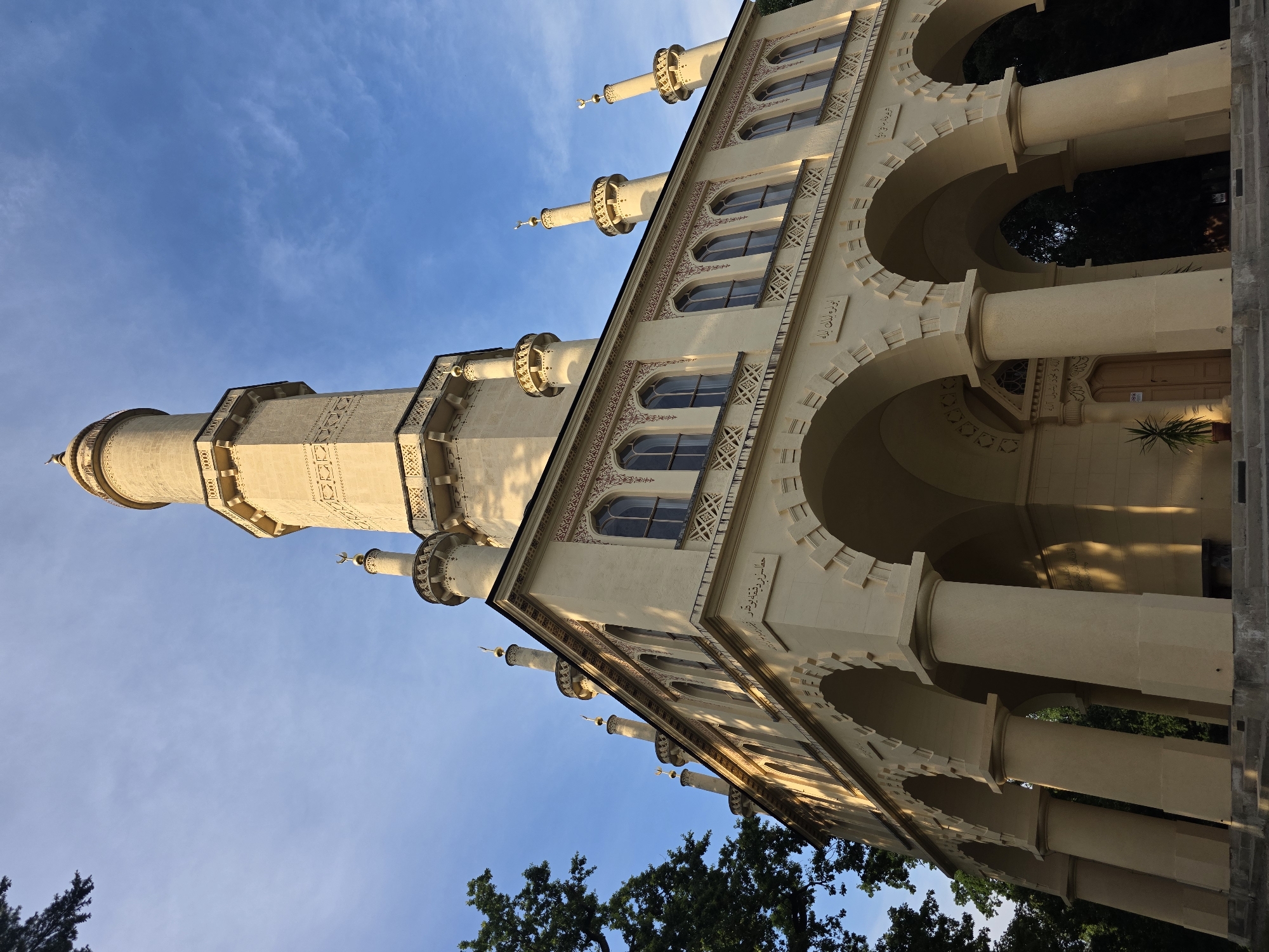 The image captures a low-angle, exterior view of the Lednice Minaret, a prominent landmark located in Lednice, Czechia. The minaret, constructed from a pale yellow material, is bathed in the warm, golden light of what appears to be late afternoon or early morning. Its architectural style is distinctively Moorish, featuring intricate decorative elements and a slender, tapering tower that culminates in a domed cap.
The minaret is part of a larger structure that includes a long, arched portico with substantial cylindrical columns. Above the arches, the building displays a series of tall, arched windows adorned with decorative patterns. The upper section of the minaret is characterized by balconies with ornate railings and smaller decorative turrets topped with crescents. Arabic inscriptions are visible on the facade of the building, beneath the windows and above the arches.
To the left of the minaret, the dark green foliage of a large tree with dense branches partially frames the structure, contrasting with the bright sky. The sky above is a clear, bright blue with wispy white clouds. The overall impression is one of grandeur and exoticism, set against a natural backdrop. The ground in the foreground appears to be a stone or concrete walkway with steps leading up to the portico. No people are visible in the image.