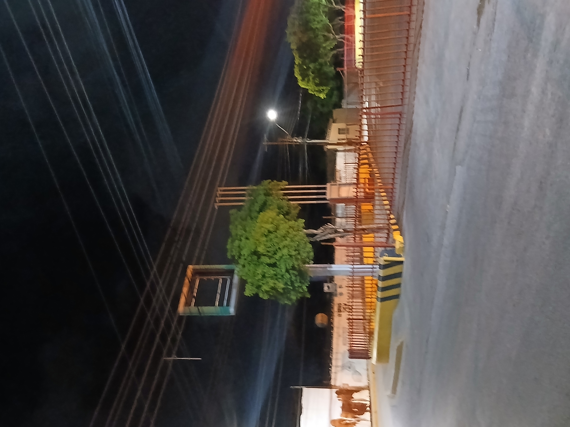 This image captures a nighttime street scene in Santa Ana, Venezuela. The foreground shows a paved road with painted yellow and black hazard stripes along the curb. Behind the curb is a fence, partially obscured by a yellow barrier. Several trees are visible, one large and green in the center, and others further back and to the right, also green. Above the trees, a street lamp with a bright light is visible on a pole. In the background, there are buildings and structures, including a large, empty, rectangular sign frame hanging above a tree. Several power lines stretch across the upper portion of the image against a dark sky. The overall impression is that of a quiet street at night.