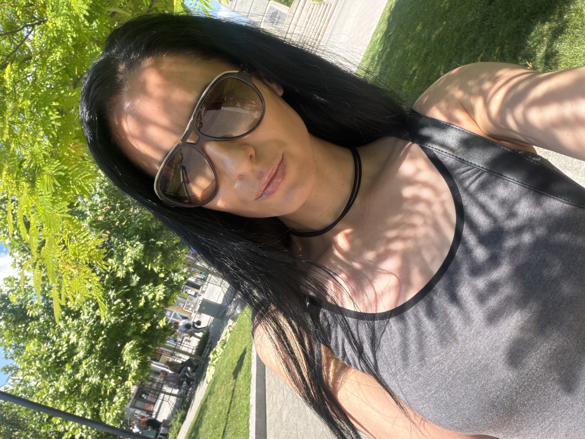 The image shows a woman with long dark hair and sunglasses, wearing a grey tank top, posing for a selfie outdoors. She is in a park with green grass and trees, with some people visible in the background. The setting appears to be a city park in Pleven, Bulgaria, possibly near the center of the city, given the urban elements and the style of the architecture in the background. The sunlight casts dappled shadows on her and the surroundings.